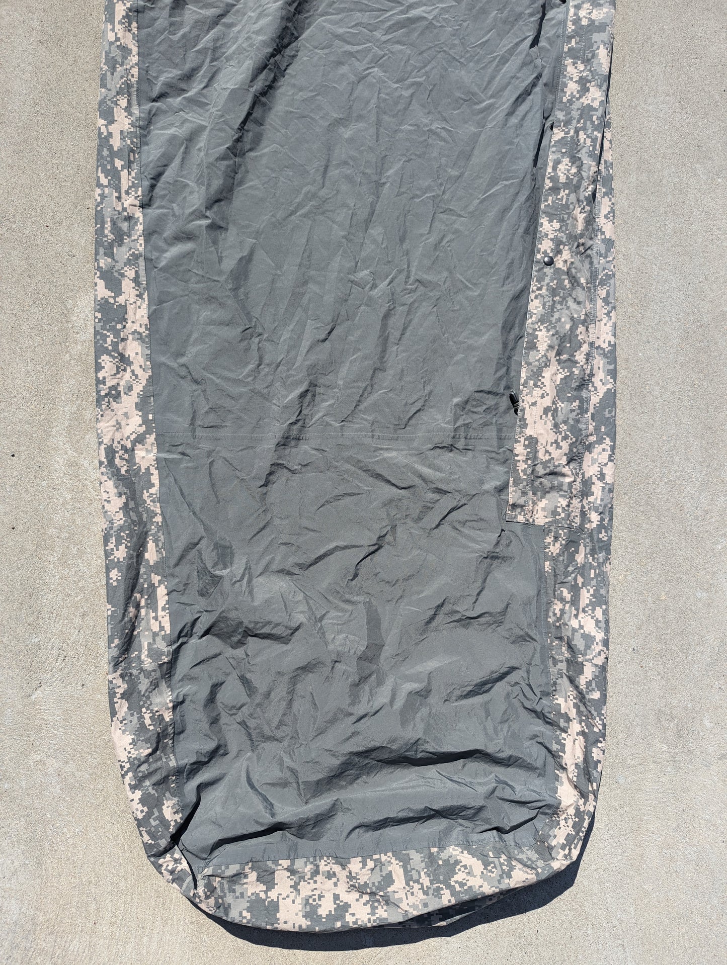 A full-length U.S. Military Surplus Modular Sleep System Bivy Cover in ACU digital camouflage, laid flat on a concrete surface, displaying the wrinkled mummy shape and waterproof fabric.