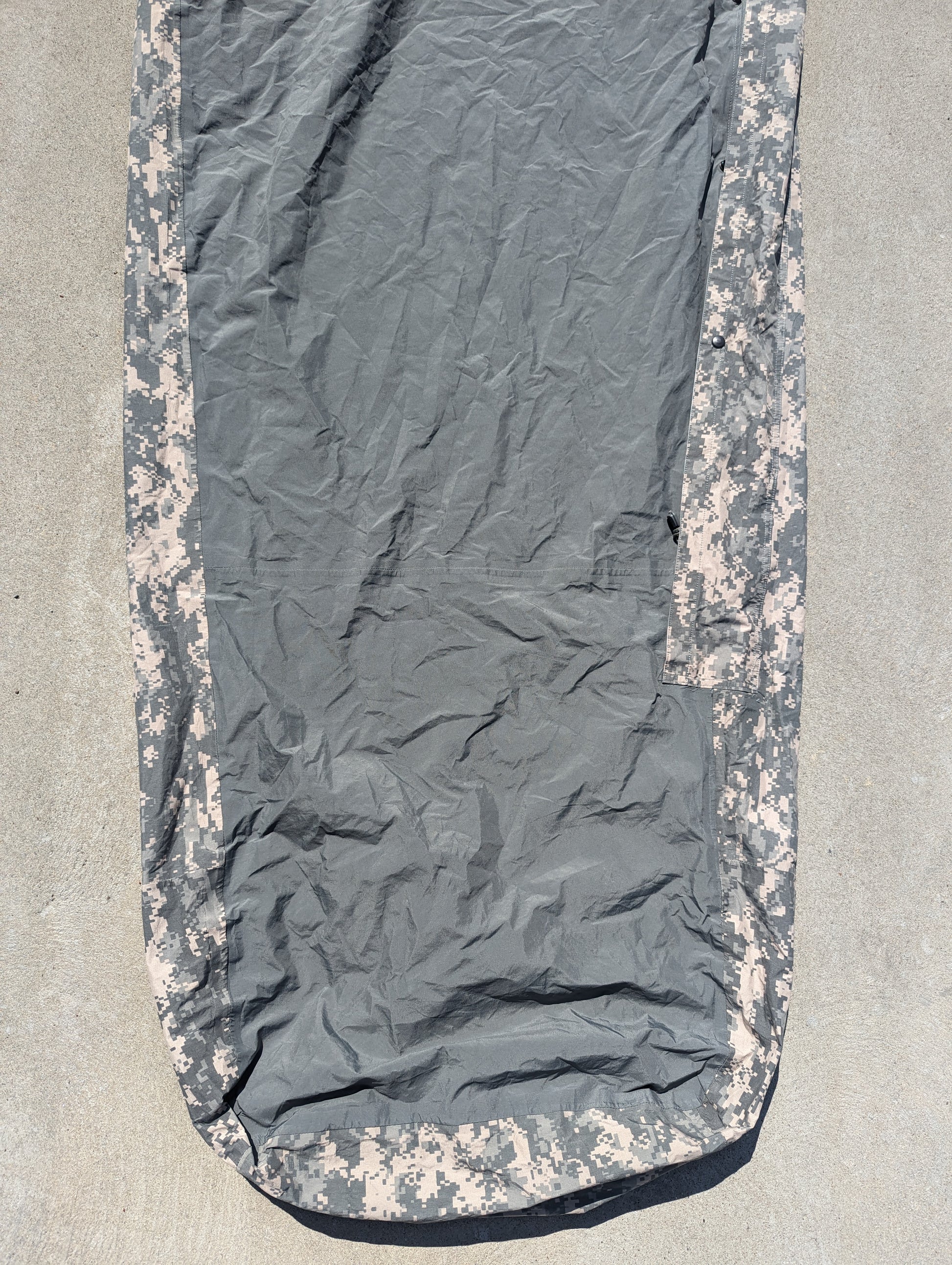 A full-length U.S. Military Surplus Modular Sleep System Bivy Cover in ACU digital camouflage, laid flat on a concrete surface, displaying the wrinkled mummy shape and waterproof fabric.