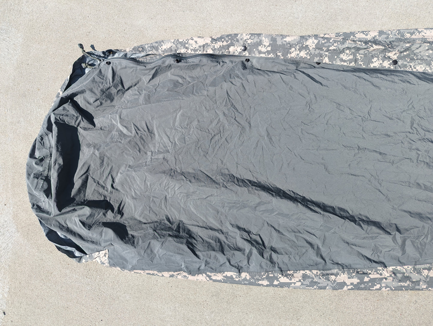 A full-length U.S. Military Surplus Modular Sleep System Bivy Cover laid flat on a concrete surface, showing the light gray waterproof material in the center and the ACU digital camouflage pattern along the hem.