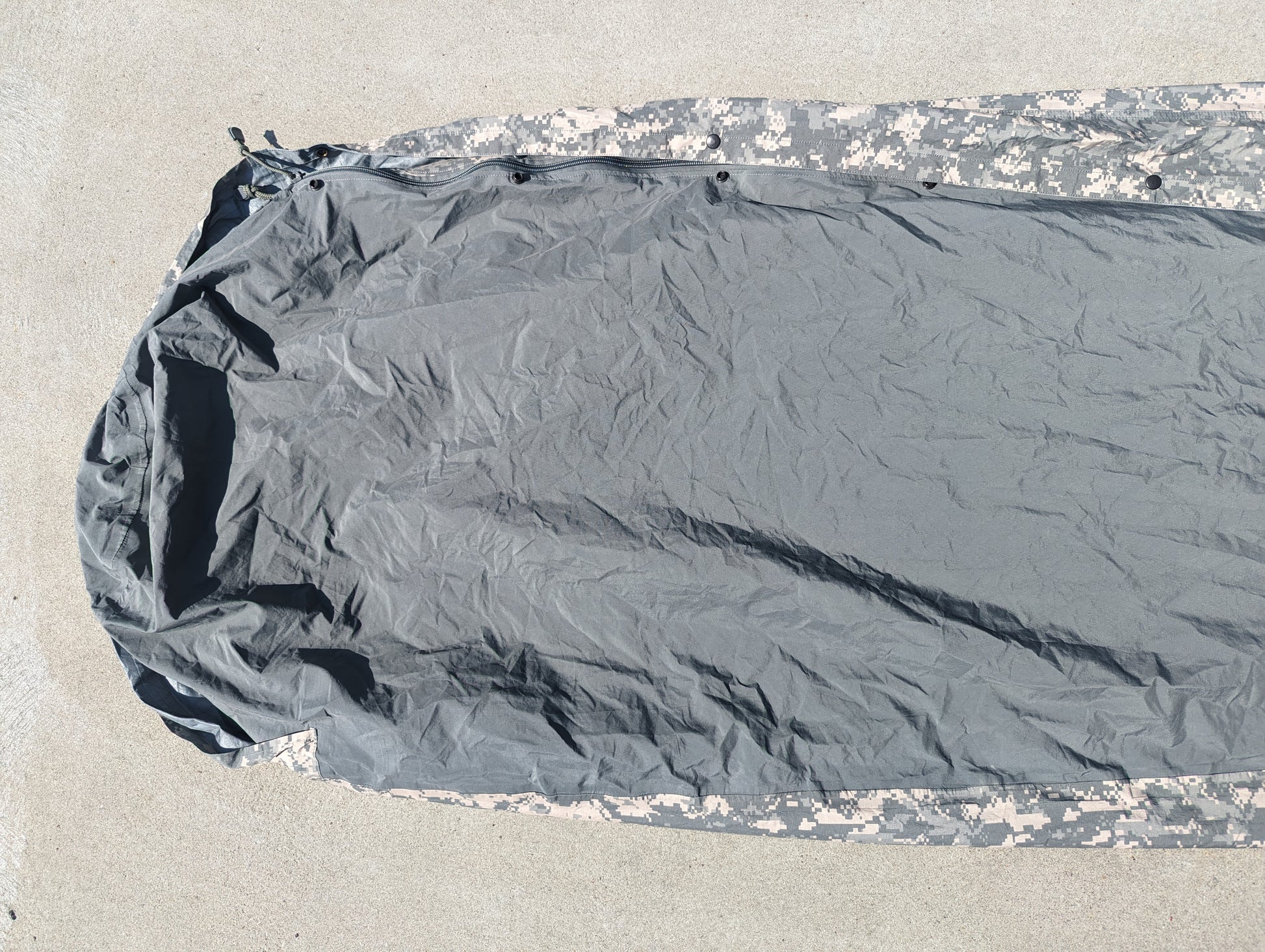 A full-length U.S. Military Surplus Modular Sleep System Bivy Cover laid flat on a concrete surface, showing the light gray waterproof material in the center and the ACU digital camouflage pattern along the hem.