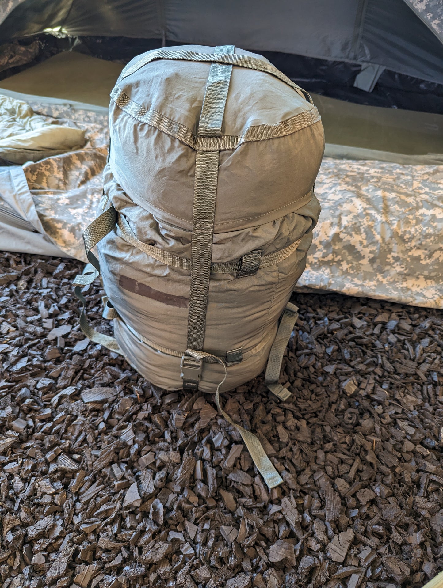 A fully compressed U.S. Military Modular Sleep System Stuff Sack in olive green, cinched tight with webbing straps and resting on a dark mulch floor next to a camouflage bivy cover.