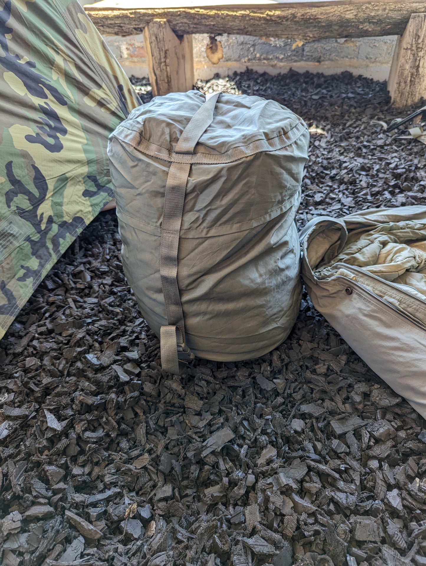 A fully compressed U.S. Military Modular Sleep System Stuff Sack in olive green, cinched tight with webbing straps and resting on a dark mulch floor next to a camouflage bivy cover.