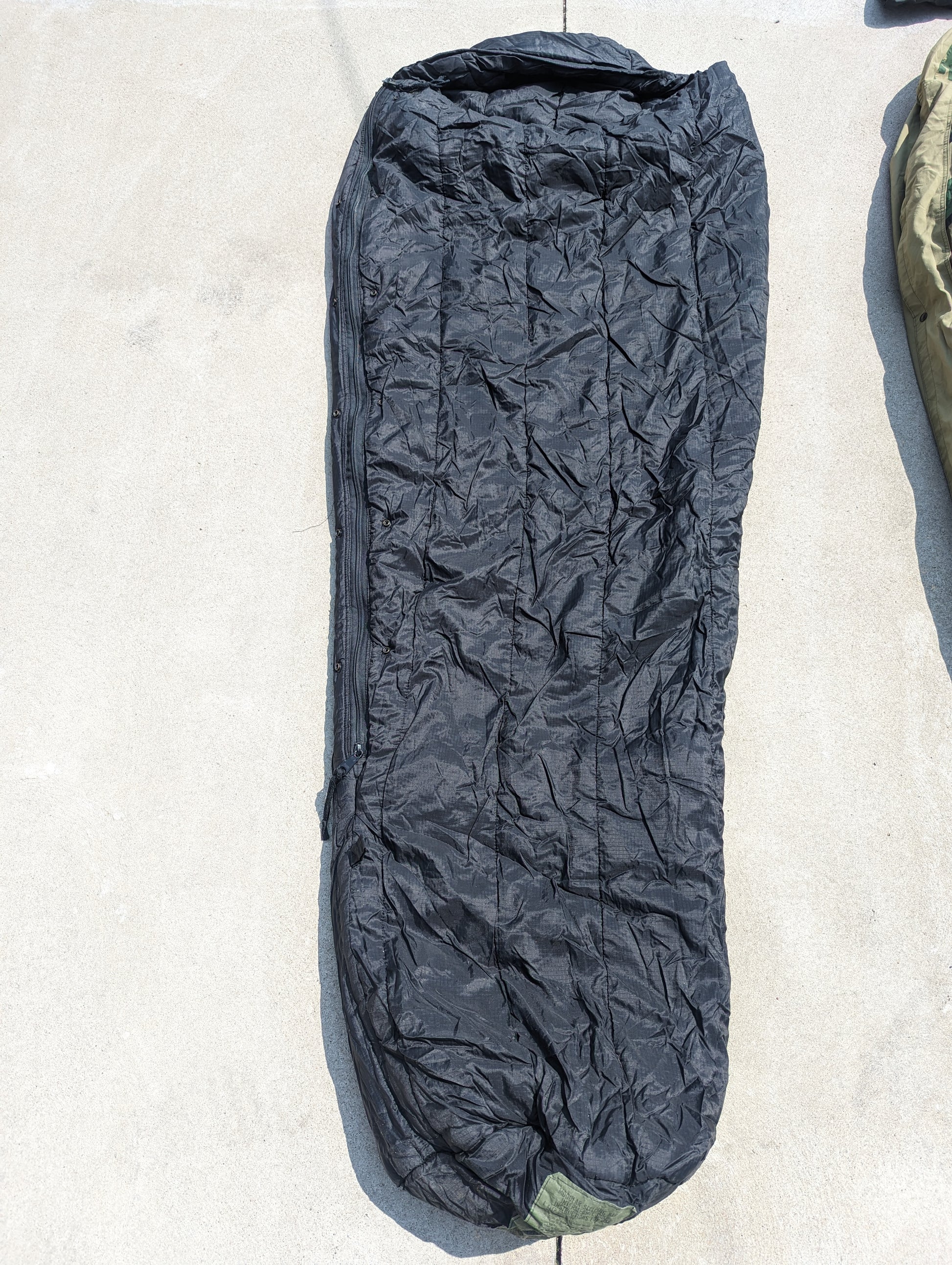 A full-length U.S. Military Modular Sleep System (MSS) Extreme Cold Weather Sleeping Bag in black, laid flat on a concrete surface, displaying its bulky, quilted, mummy shape.