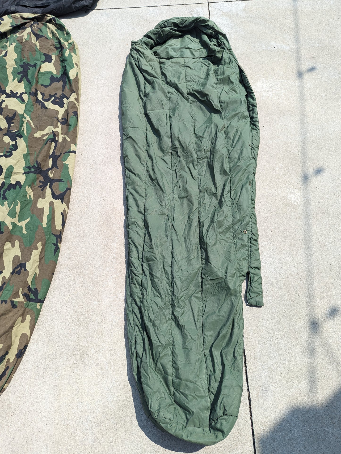 A view of two U.S. Military Modular Sleep System components laid side-by-side on concrete: one in Woodland camouflage and one in solid olive green, both zipped and displaying their mummy shape.