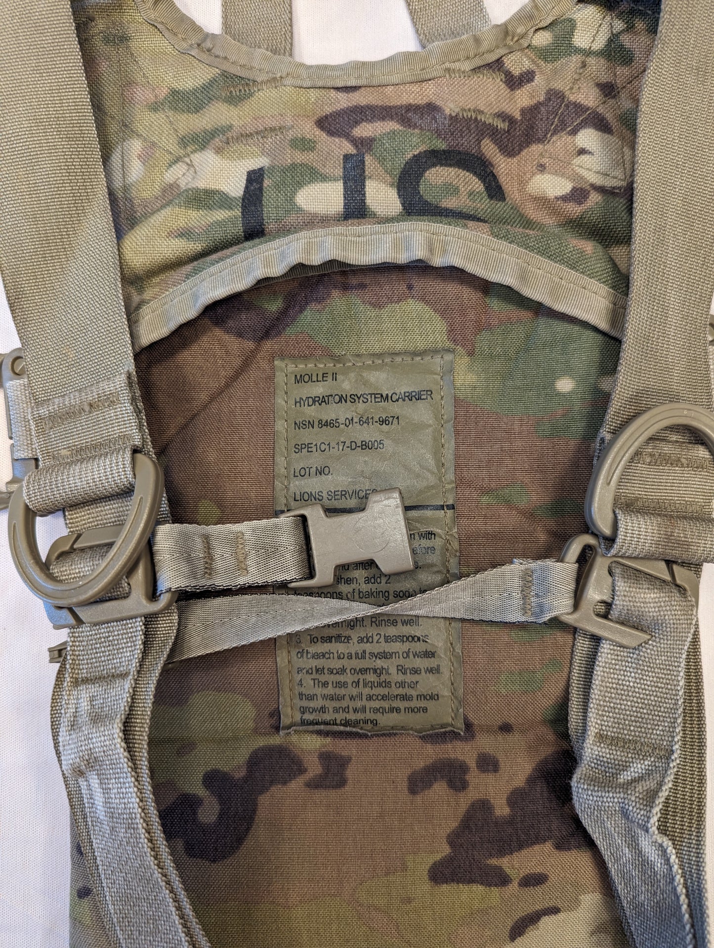 A close-up view of the back panel of a U.S. Military OCP/Multicam Hydration Carrier, showing the padded shoulder harness, tan adjustment webbing, and a centrally located military issue tag.