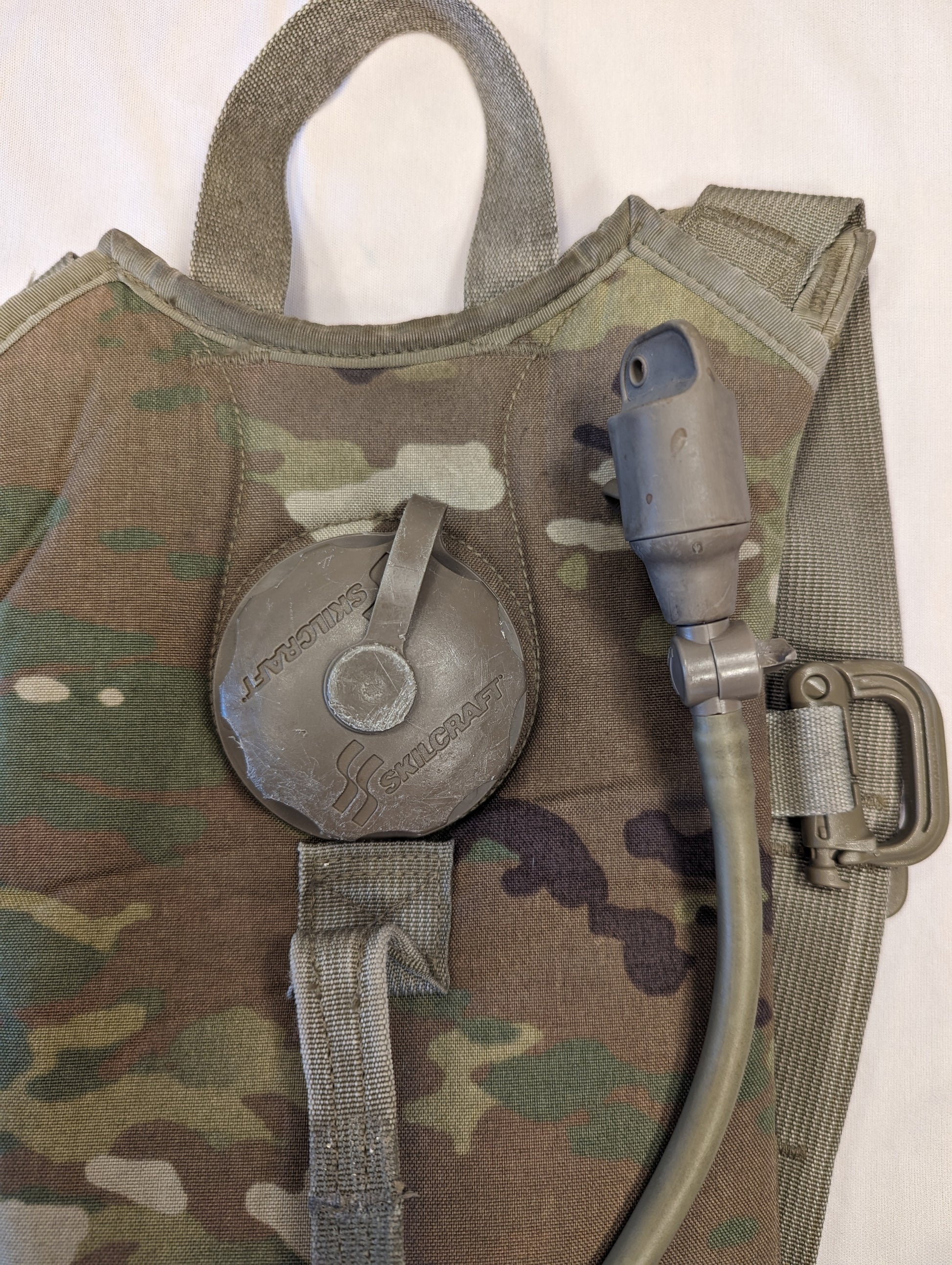 A close-up view of the filler port on a used OCP/Multicam Hydration Carrier, featuring the large plastic cap, the attached insulated drinking hose, and a plastic D-ring attachment.