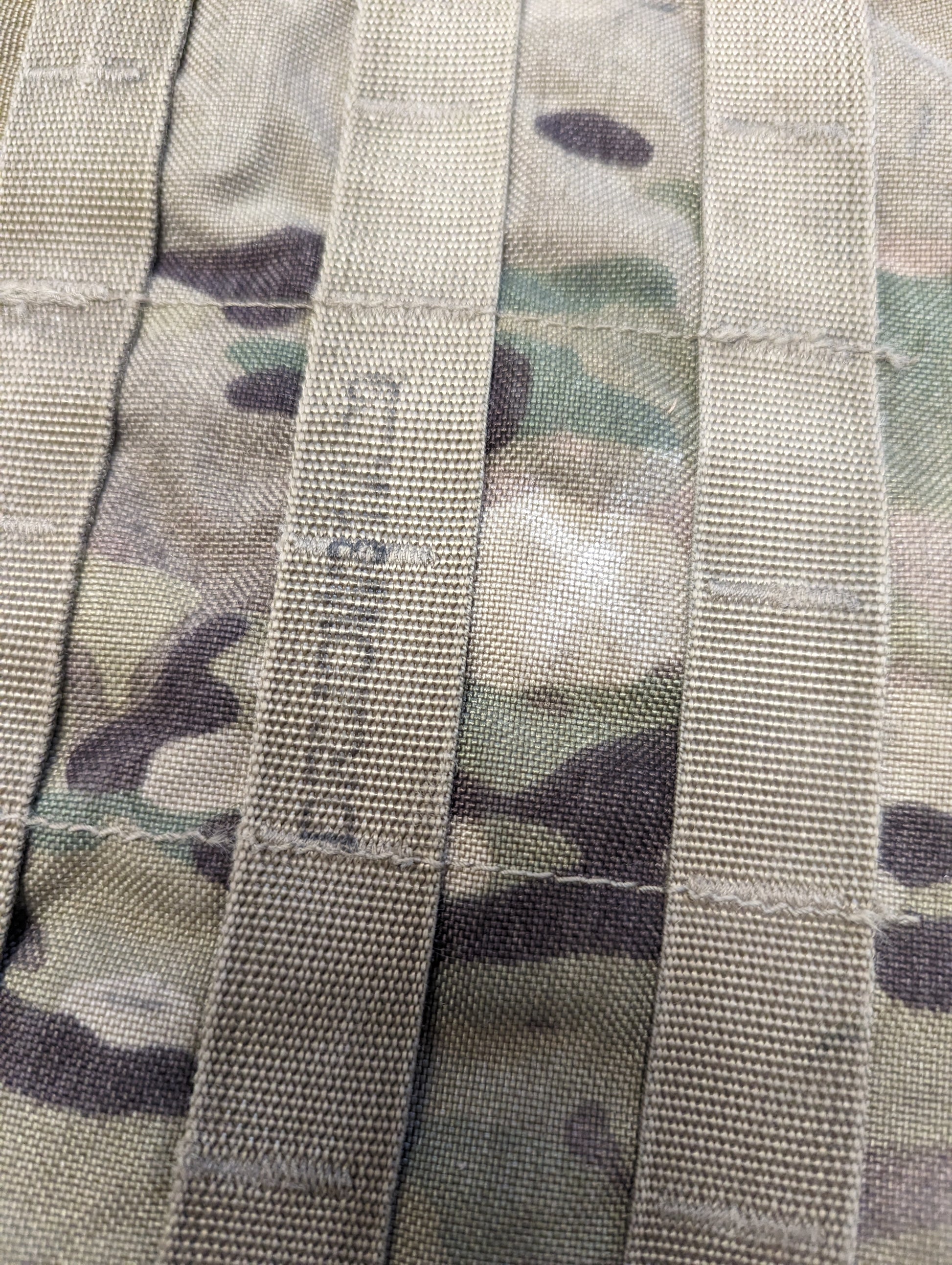 A close-up detail shot of OCP/Multicam camouflage PALS/MOLLE webbing, showing the horizontal and vertical strap construction, with the letters 'US' faintly marked on one of the straps.
