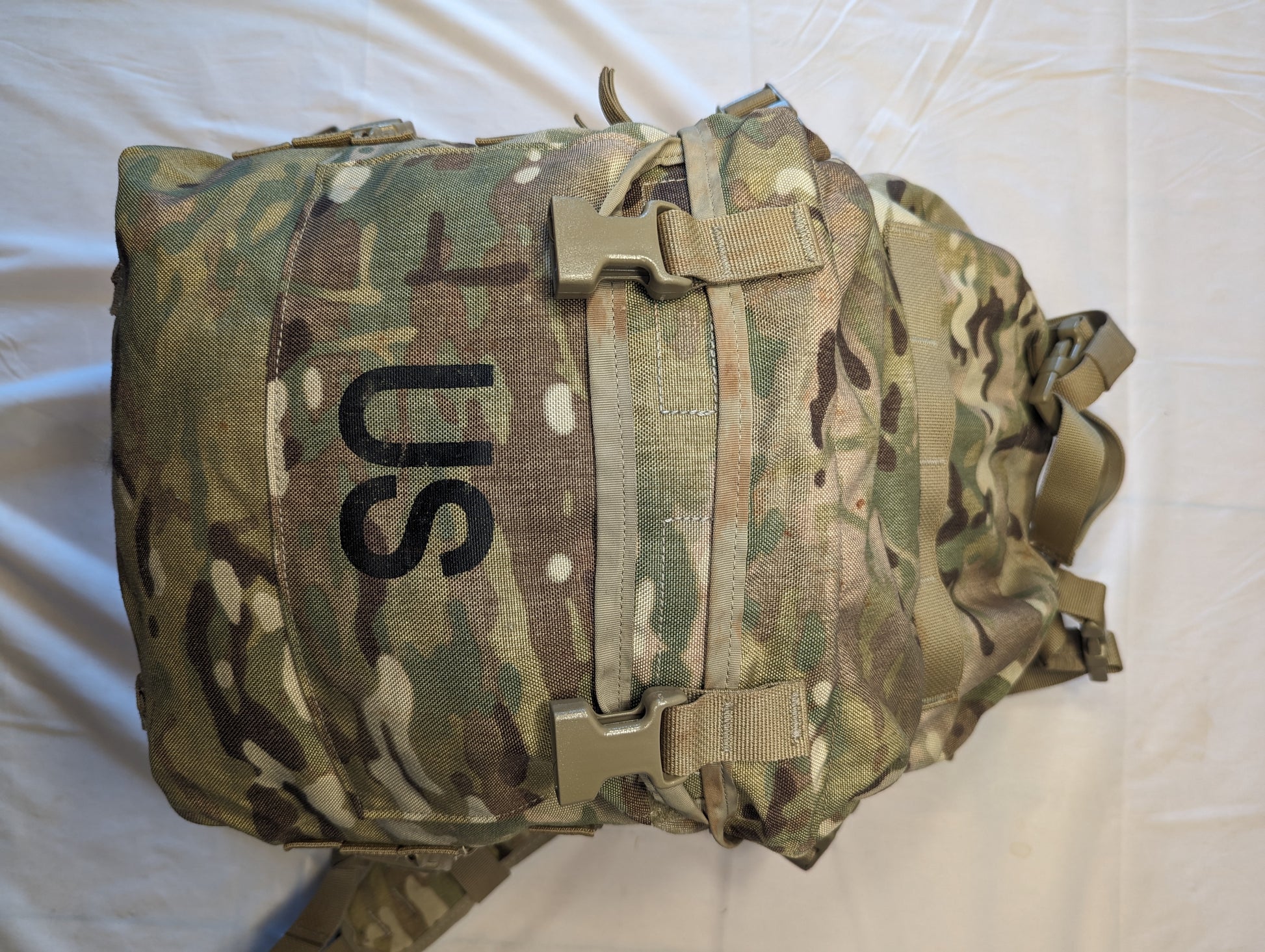 A close-up front view of a U.S. Military Assault Pack in OCP/Multicam camouflage, featuring a large stenciled 'US' mark and two tan quick-release buckles securing the top flap.