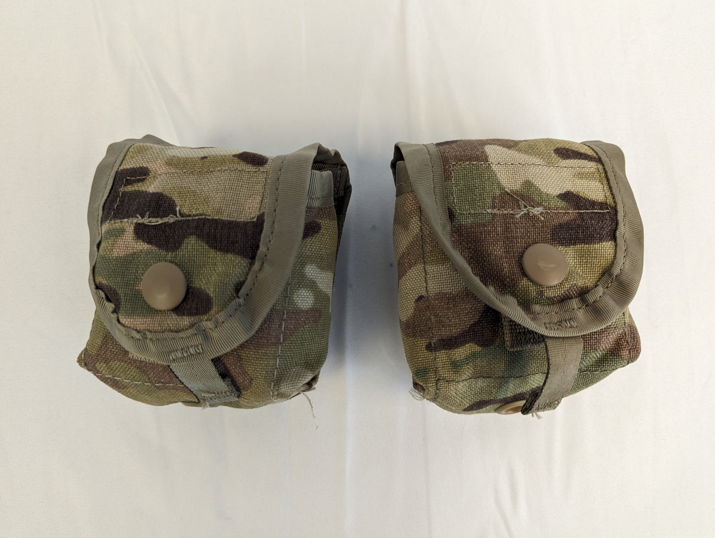 A studio shot of two U.S. Military Surplus hand grenade pouches in OCP/Multicam camouflage, shown side-by-side with closed snap-button flaps on a wrinkled white background.
