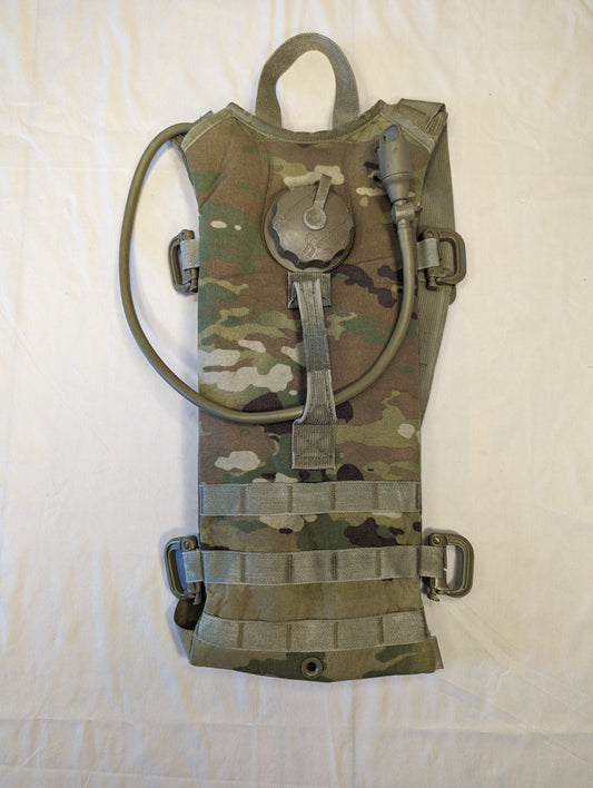 A used U.S. Military Hydration Carrier in OCP/Multicam camouflage, laid flat, featuring a visible black filler cap, drinking hose, and horizontal MOLLE webbing, displaying a worn texture.