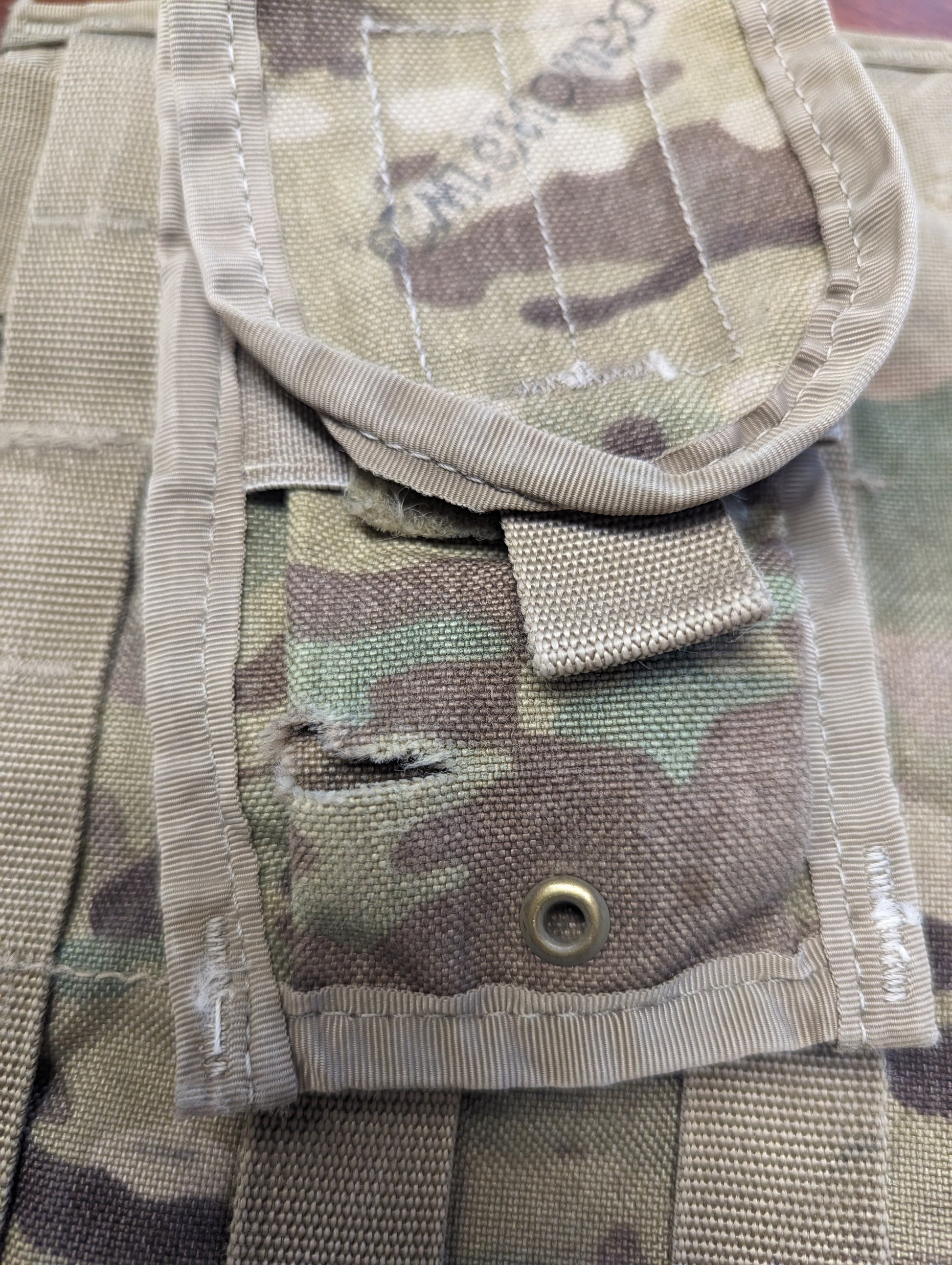 An extreme close-up detail shot of an OCP/Multicam tactical pouch flap, highlighting a tear or hole in the fabric just above a brass drainage grommet, documenting a specific surplus defect.