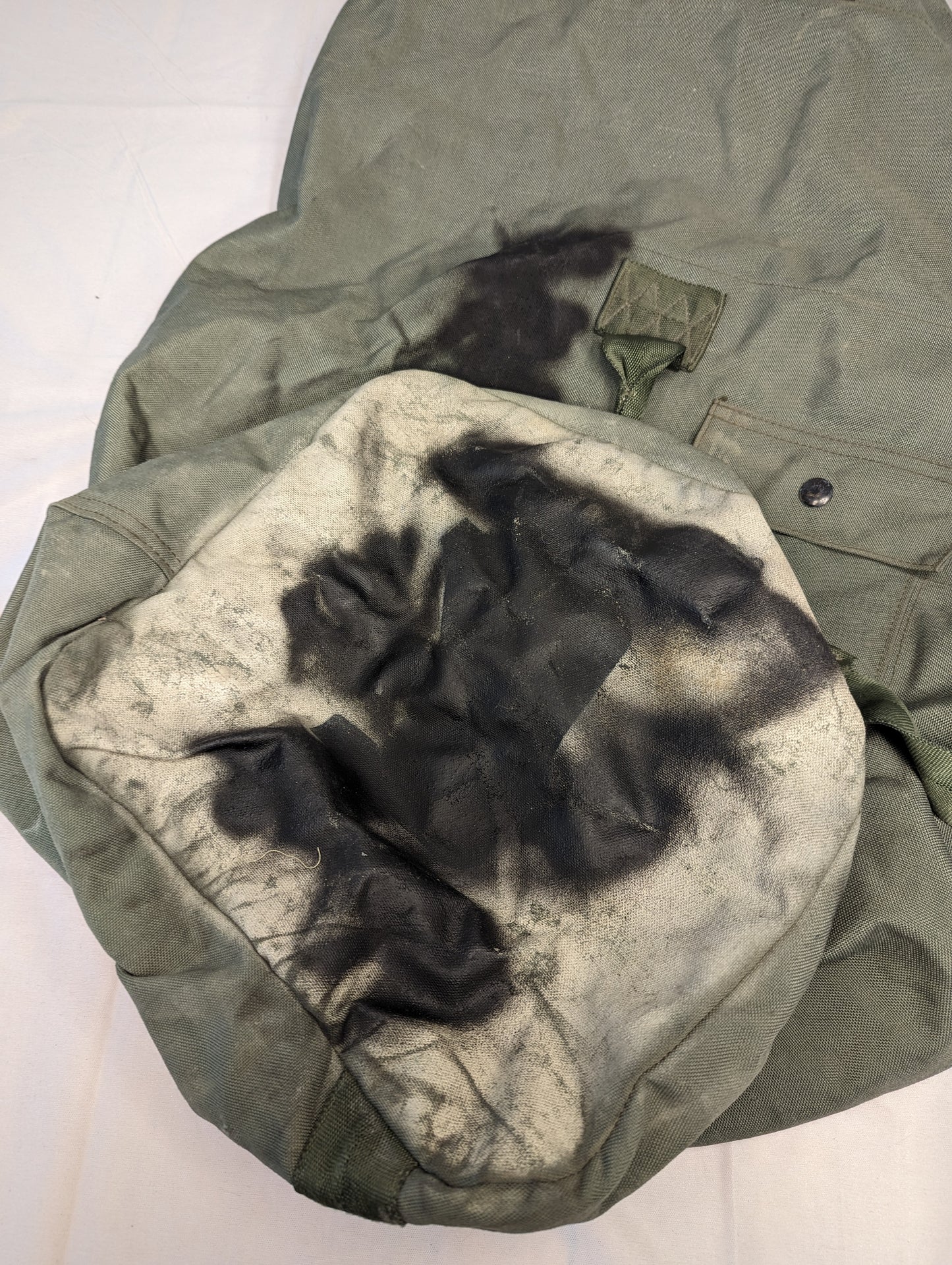 An extreme close-up photograph of dark olive green quilted military fabric, showing a large, severe black soot stain and discoloration, documenting a major defect.