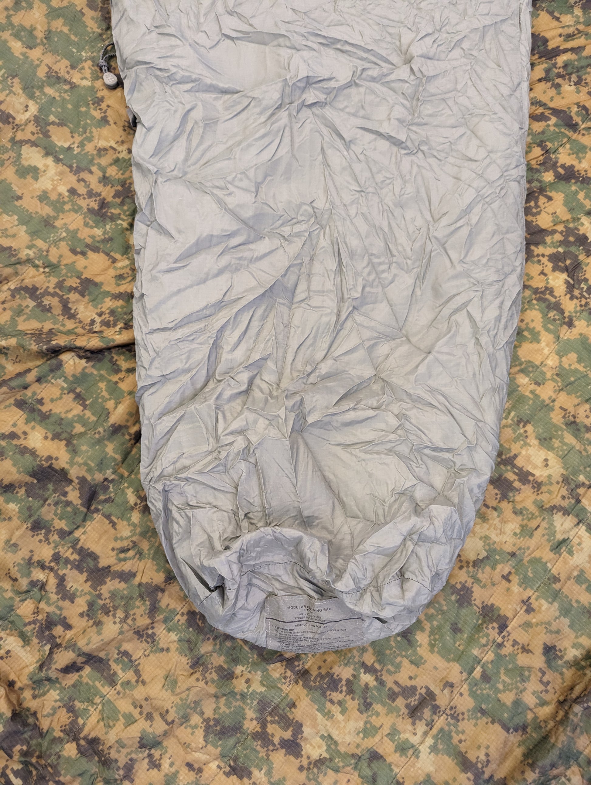 A close-up overhead view of a U.S. Military Surplus Patrol Sleeping Bag in wrinkled foliage green, with the hood cinched closed, resting on a camouflage fabric background.