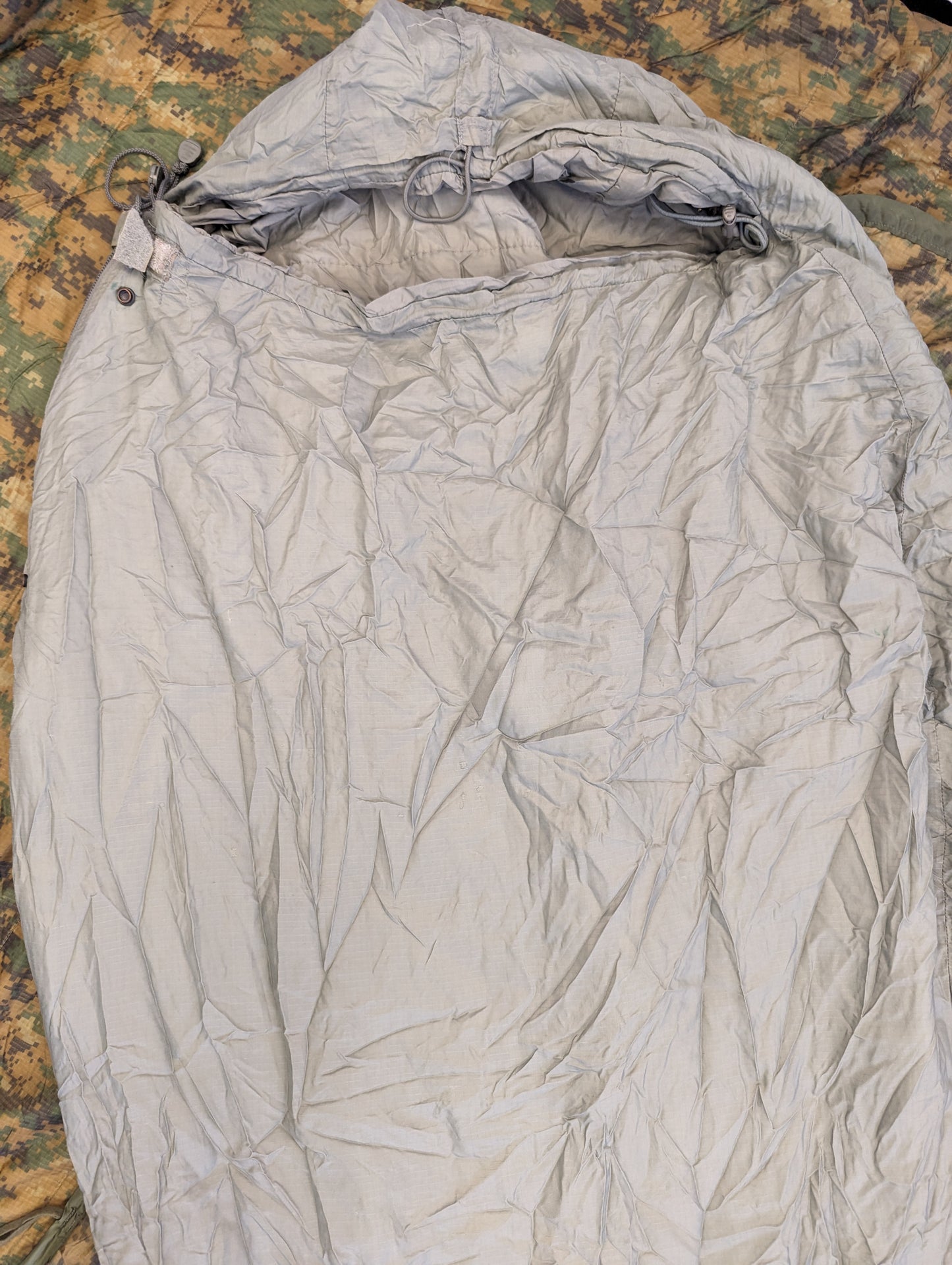 A close-up overhead view of a U.S. Military Surplus Patrol Sleeping Bag in wrinkled foliage green, with the hood cinched closed, resting on a camouflage fabric background.