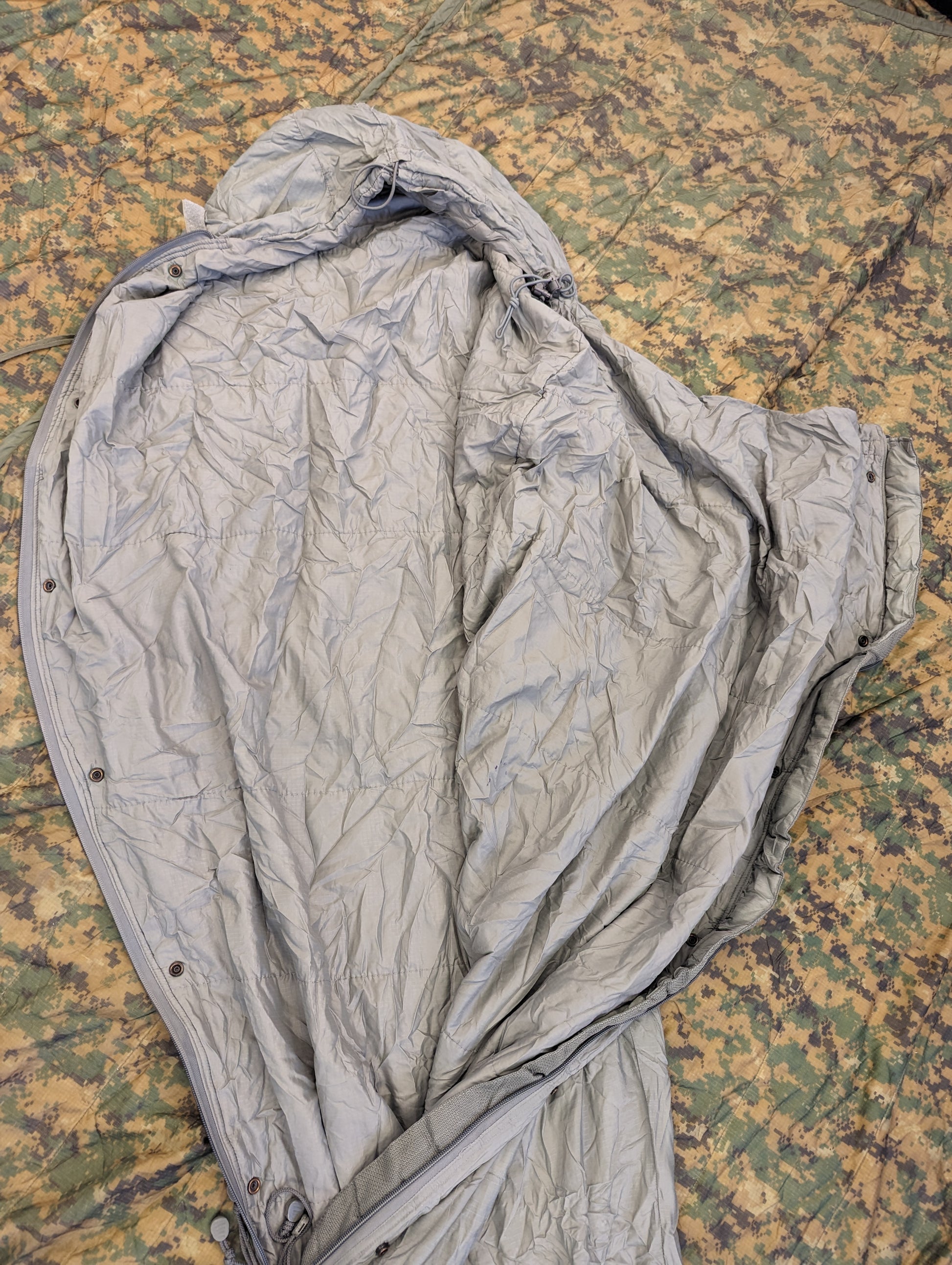 A close-up overhead view of a U.S. Military Surplus Patrol Sleeping Bag in wrinkled foliage green, with the hood cinched closed, resting on a camouflage fabric background.