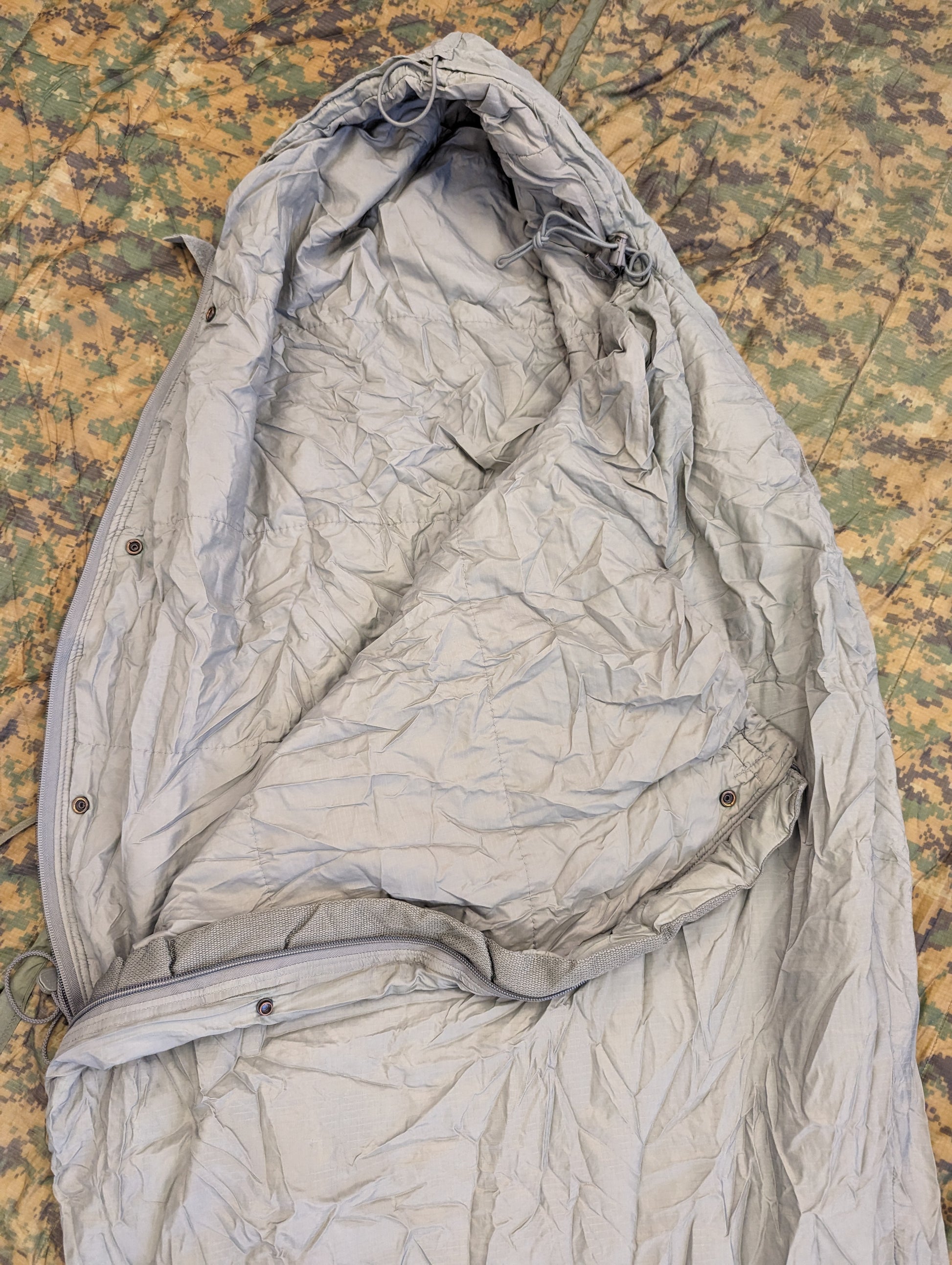 A close-up overhead view of a U.S. Military Surplus Patrol Sleeping Bag in wrinkled foliage green, with the hood cinched closed and snaps visible, resting on a camouflage fabric background.