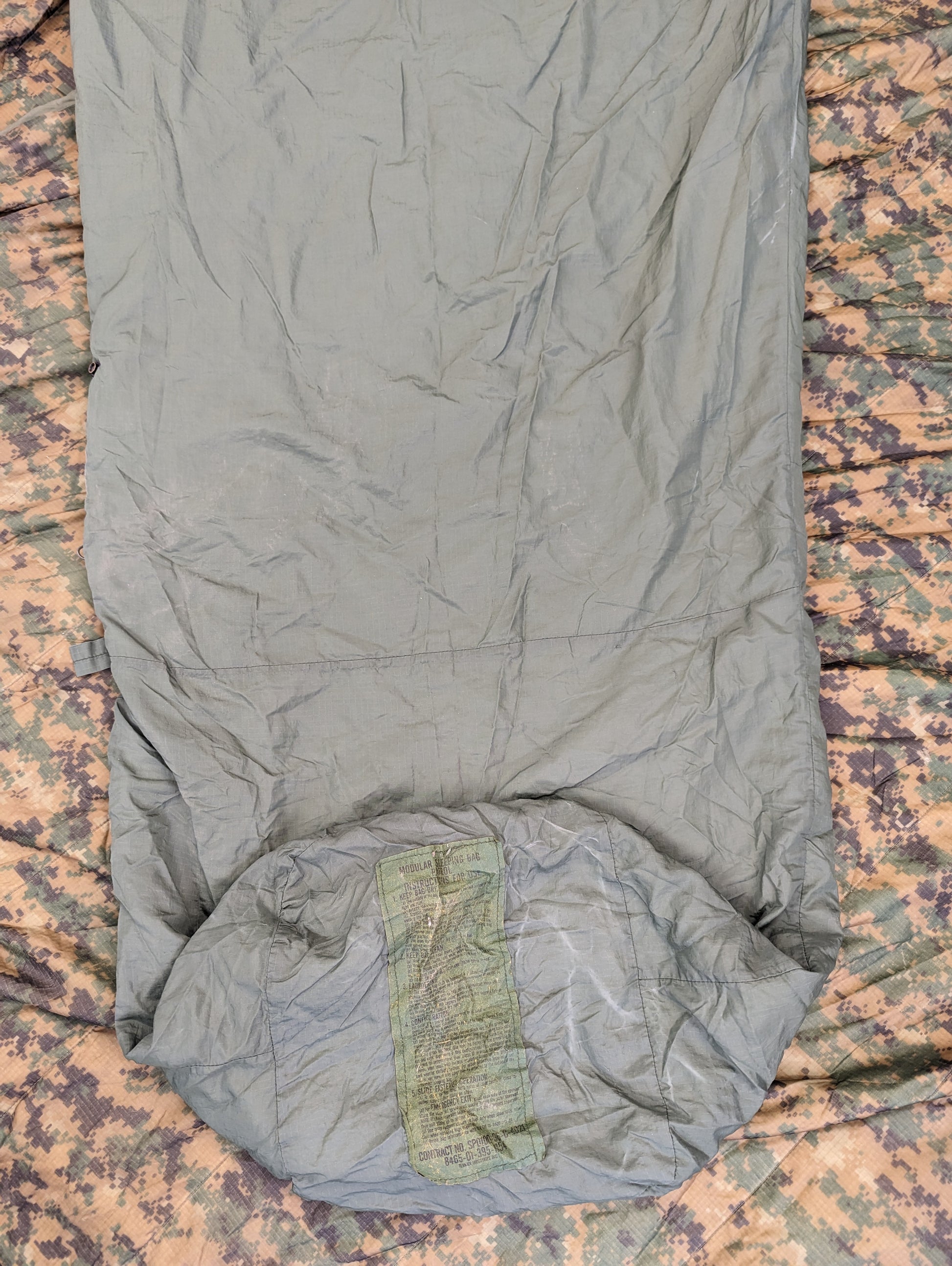 A full-length U.S. Military Surplus Patrol Sleeping Bag in light olive green, unrolled and laid vertically on a dark camouflage fabric background, showing the hood and foot box.