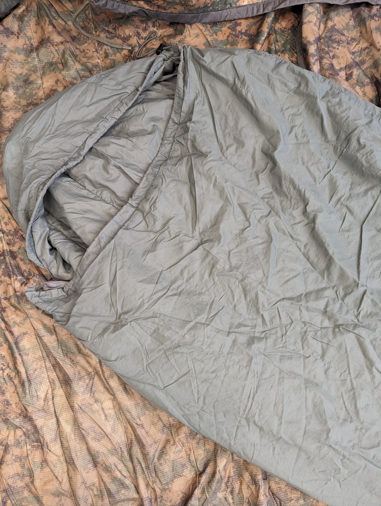 A close-up view of the top half of a U.S. Military Surplus Patrol Sleeping Bag in light olive green, resting on a dark camouflage background.