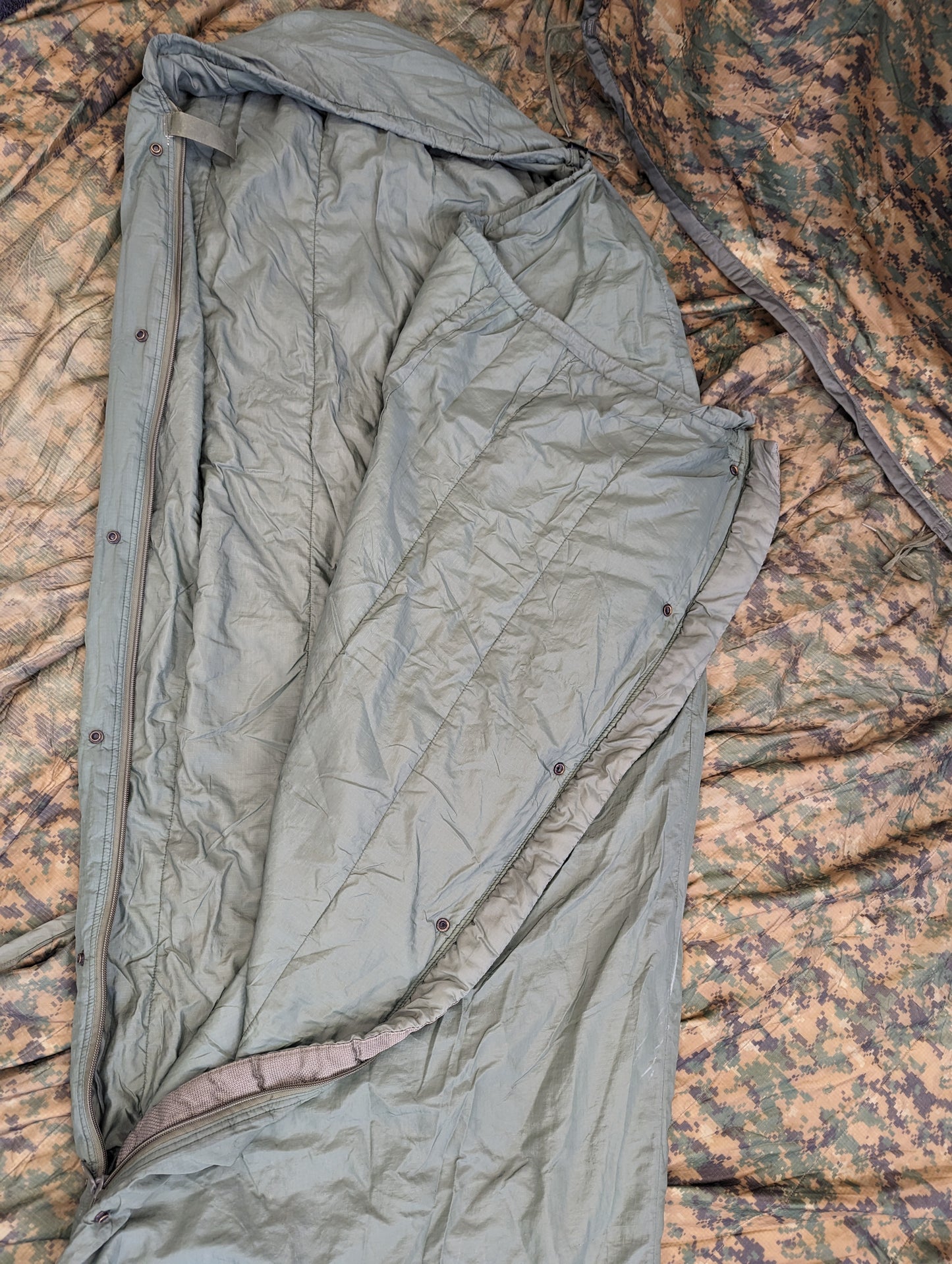 A close-up overhead view of a U.S. Military Surplus Patrol Sleeping Bag in light olive green, fully zipped with the hood cinched, resting on a camouflage fabric background.