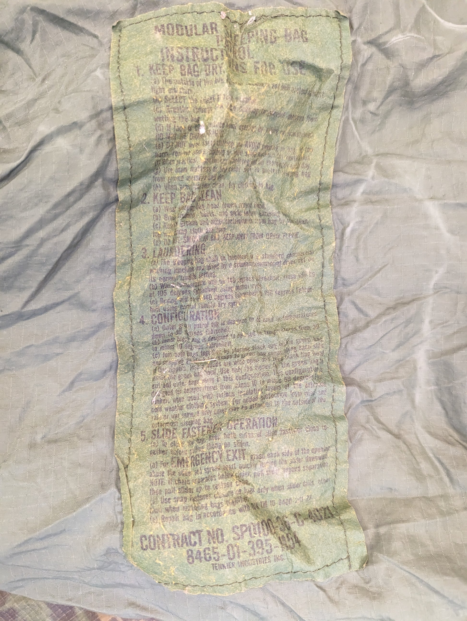 A close-up photograph of the original U.S. Military issue tag sewn into the Patrol Sleeping Bag, clearly displaying the product name, contract number, and Defense Logistics Agency information.