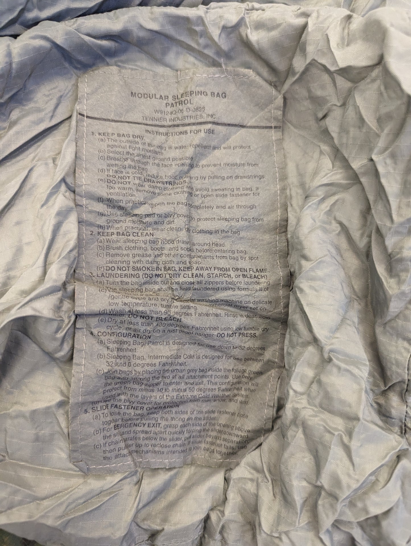 A close-up photograph of the original U.S. Military issue tag sewn into the Patrol Sleeping Bag, displaying the product name, 'MODULAR SLEEPING BAG: PATROL', and detailed laundering instructions.