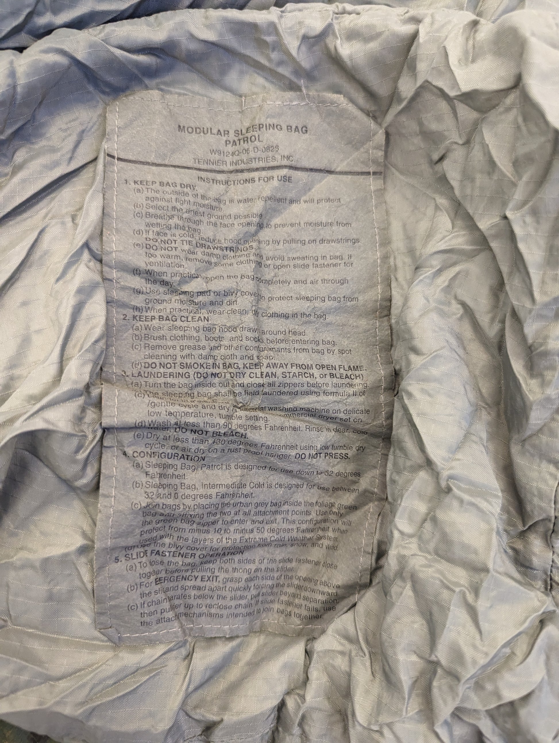 A close-up photograph of the original U.S. Military issue tag sewn into the Patrol Sleeping Bag, displaying the product name, 'MODULAR SLEEPING BAG: PATROL', and detailed laundering instructions.