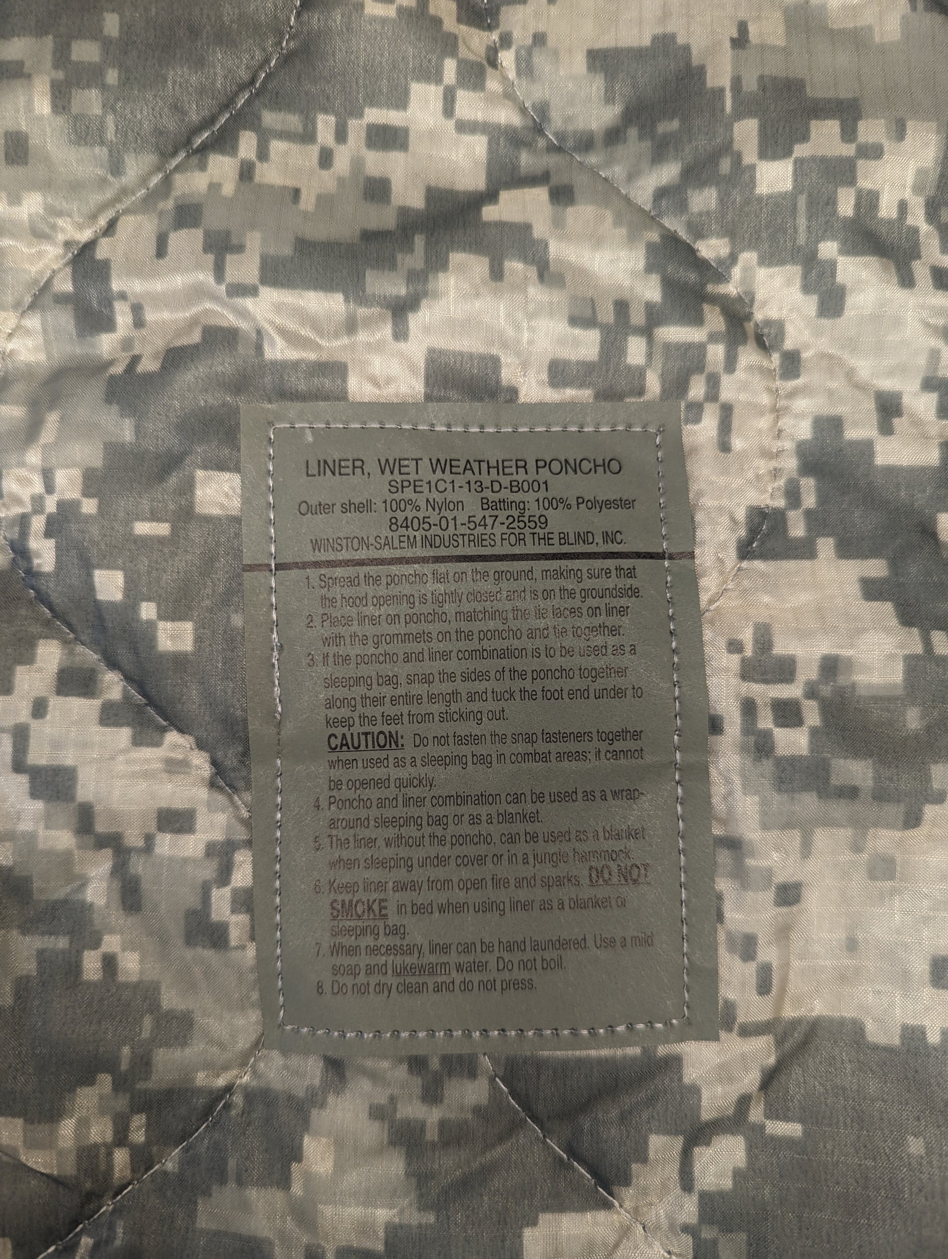 A close-up photograph of the official U.S. Military issue and care tag sewn into the ACU digital camouflage Poncho Liner, clearly displaying 'LINER, WET WEATHER PONCHO', NSN 8415-01-547-2559, and detailed caution and usage instructions.
