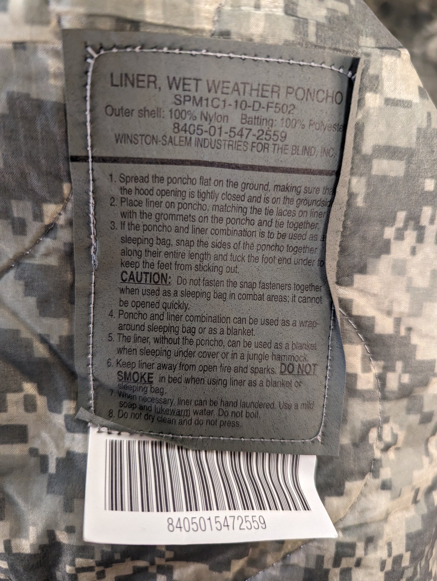 A close-up photograph of the official U.S. Military issue and care tag sewn into the ACU digital camouflage Poncho Liner, clearly displaying 'LINER, WET WEATHER PONCHO', NSN 8405-01-547-2559, and detailed usage and safety instructions.