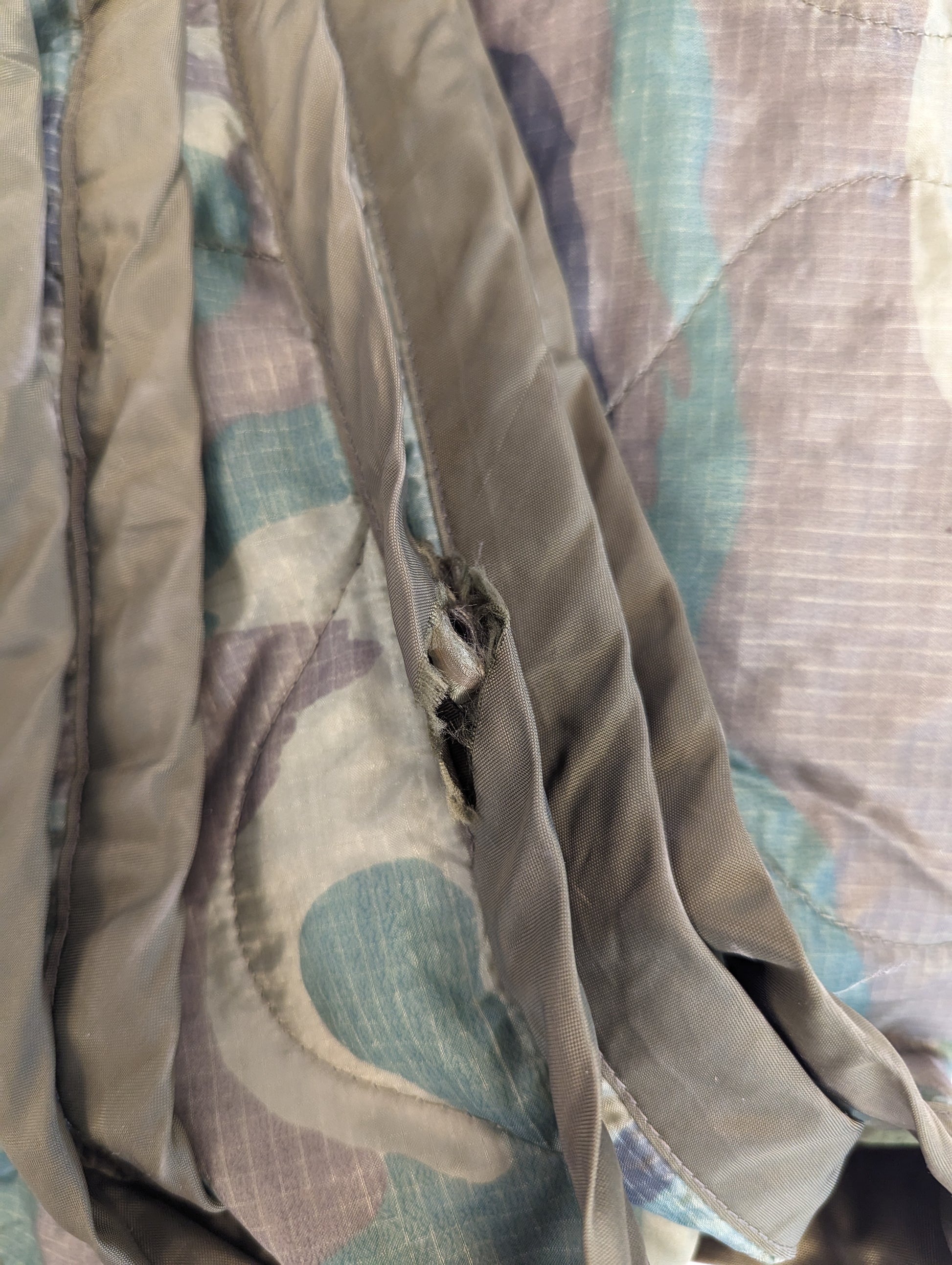 An extreme close-up photograph of a U.S. Military Poncho Liner in camouflage, showing a significant tear, rip, or hole in the quilted fabric near the binding, documenting a defect.