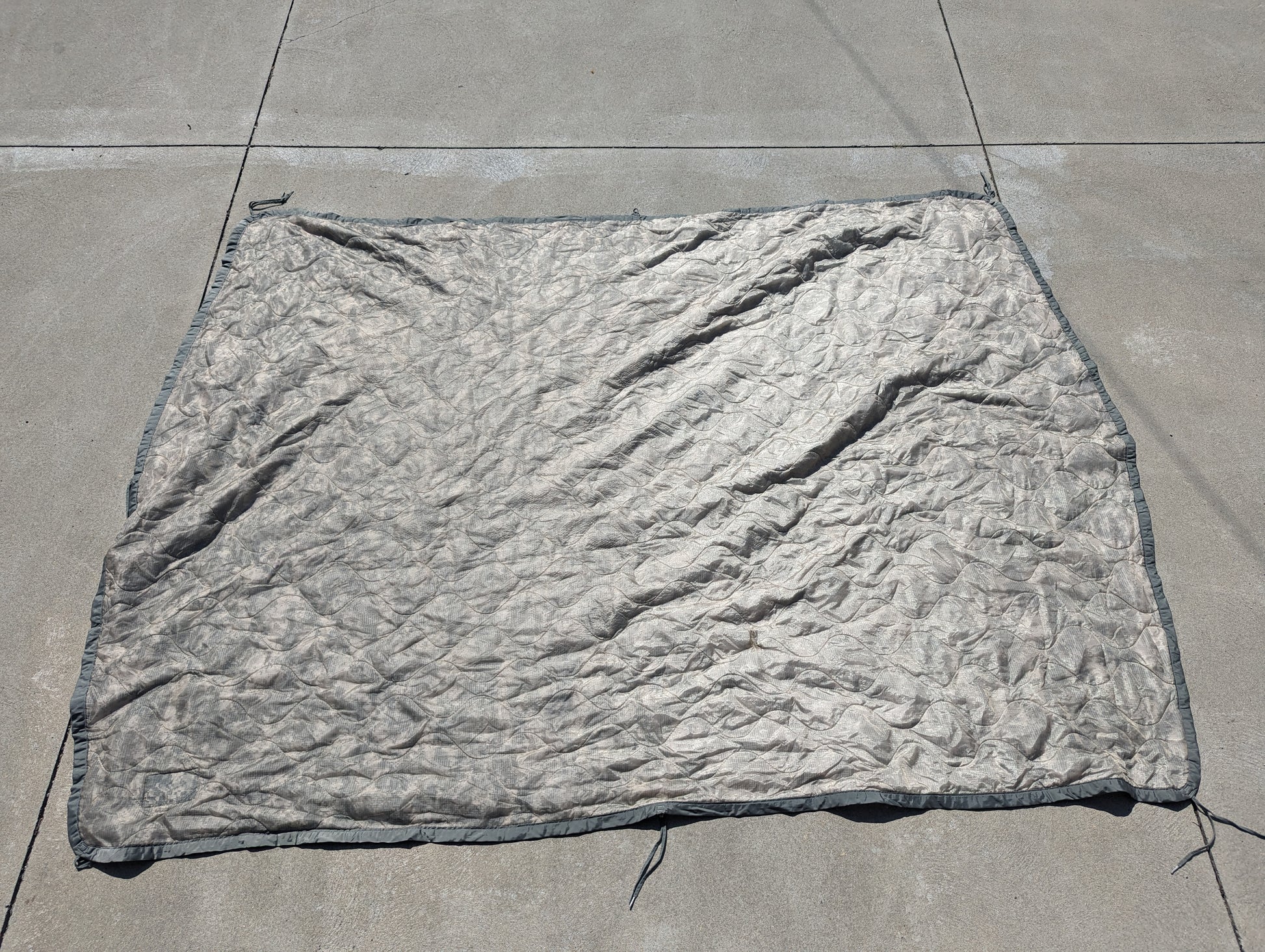 A full-length U.S. Military Surplus Poncho Liner (Woobie) in a muted gray/ACU camouflage, laid flat on a concrete surface, displaying its wrinkled texture, edge binding, and attachment tie cords.