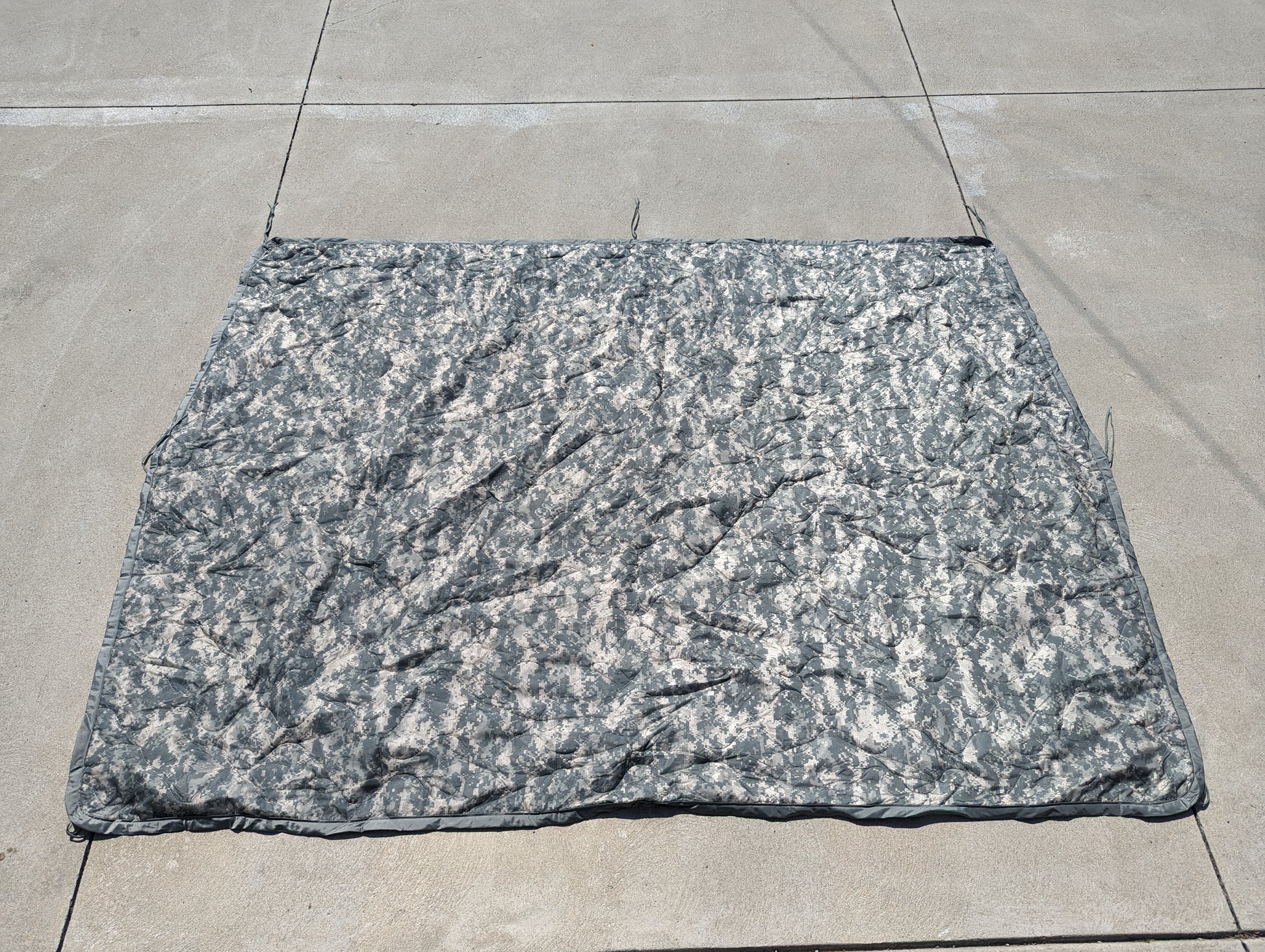 A U.S. Military Surplus Poncho Liner (Woobie) in ACU digital camouflage, fully unfolded and laid flat on a concrete surface, displaying its full rectangular size and wrinkled, quilted texture.