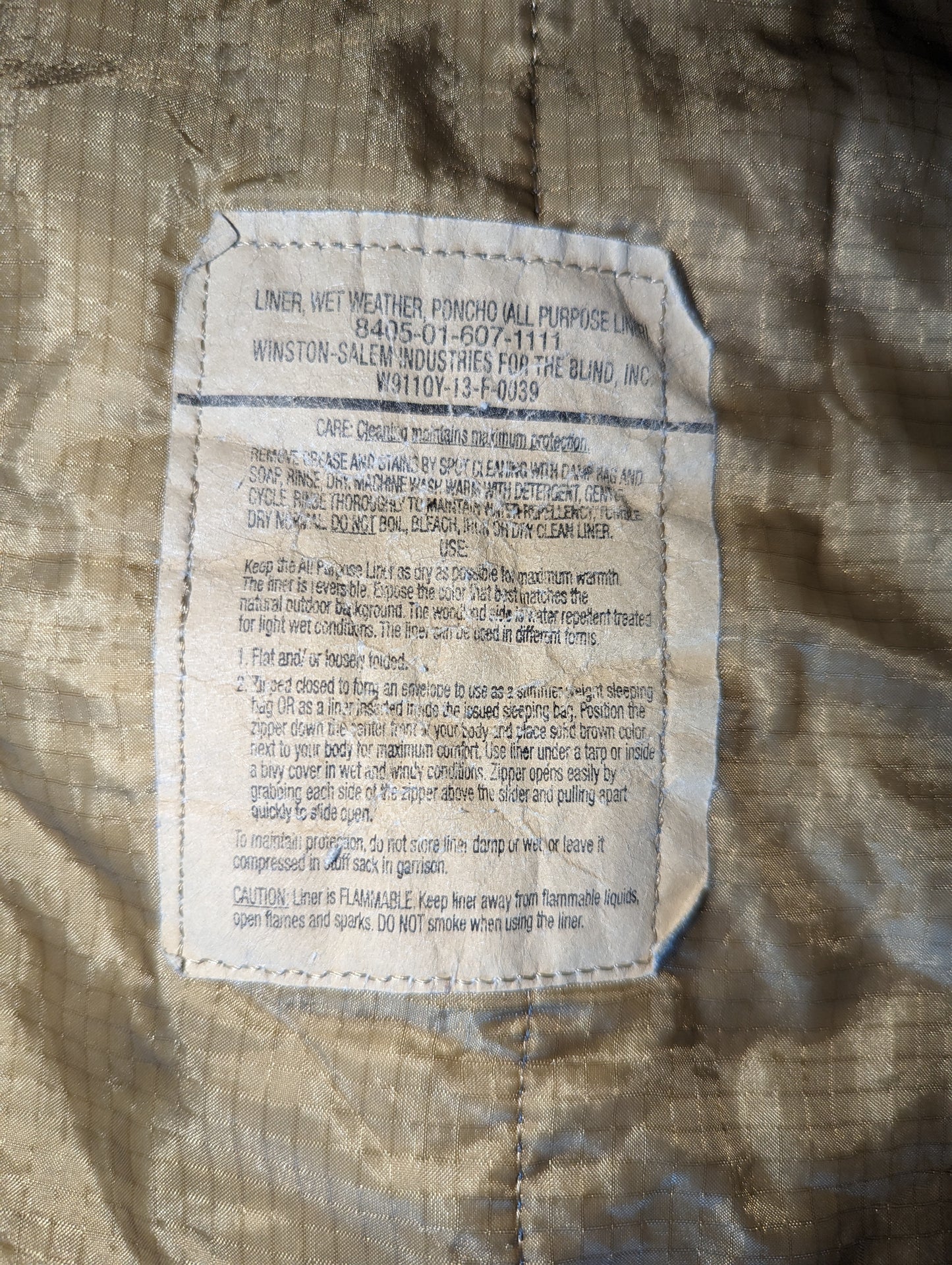 A close-up photograph of the official U.S. Military issue and care tag sewn into the tan Poncho Liner (Woobie), displaying the product name, NSN, and detailed laundering instructions.