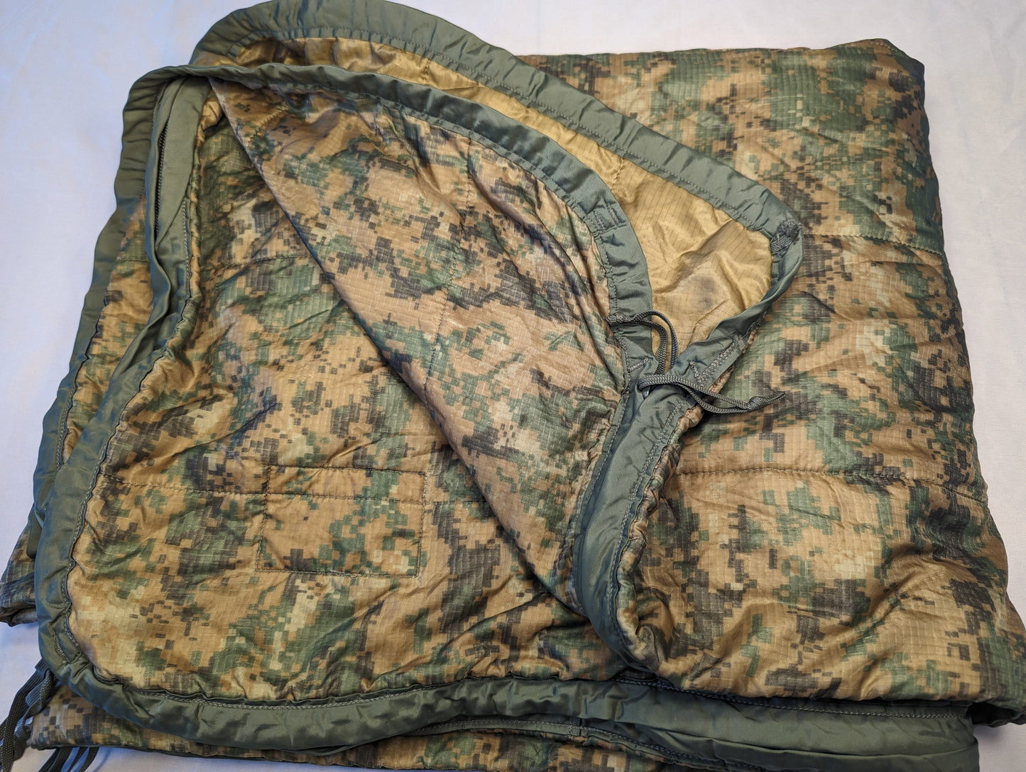 A U.S. Military Surplus Poncho Liner (Woobie) in MARPAT digital camouflage, laid flat and folded, displaying the quilted pattern, olive drab edge binding, and the tan inner lining.