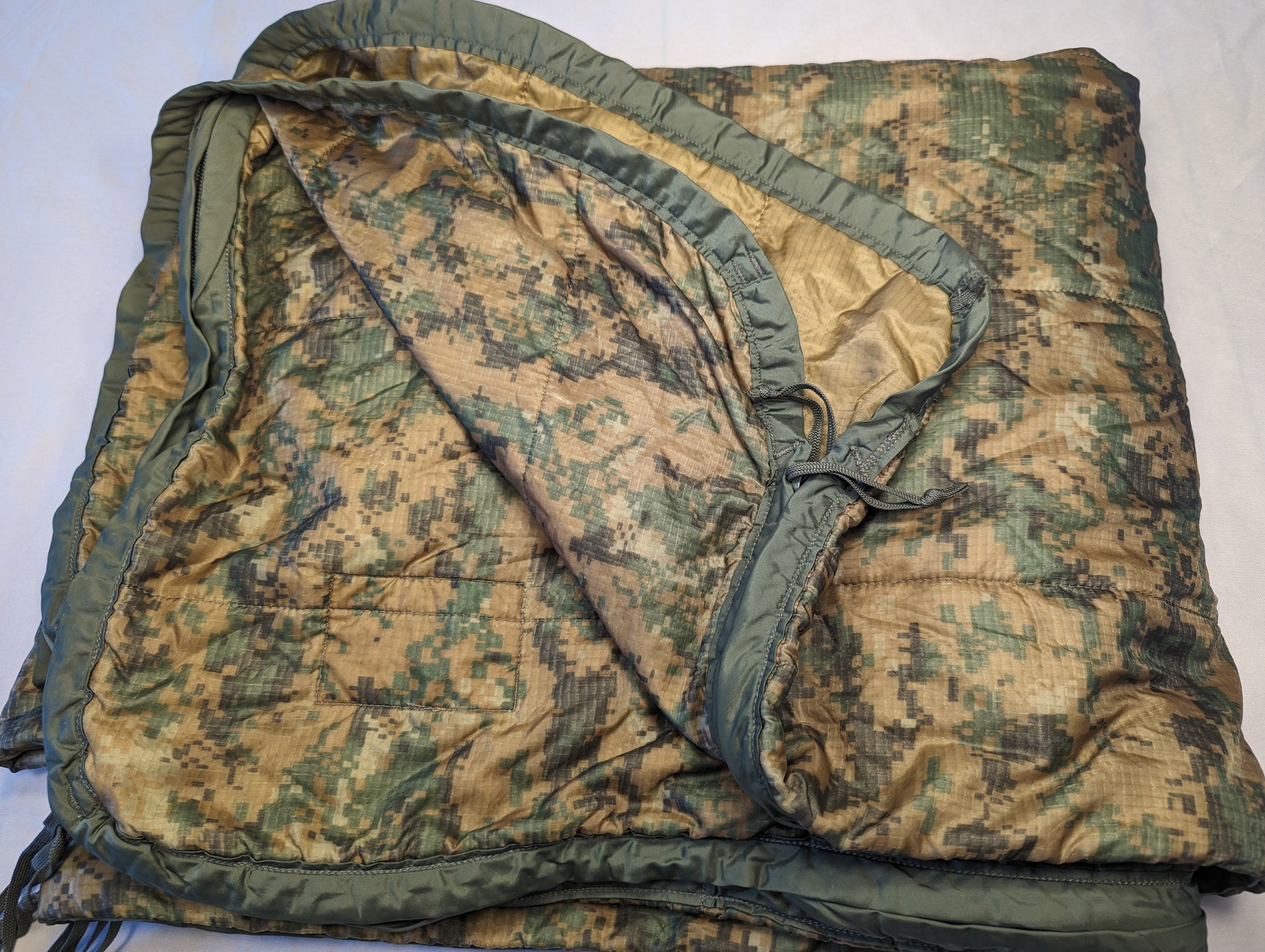A U.S. Military Surplus Poncho Liner (Woobie) in MARPAT digital camouflage, laid flat and folded, displaying the quilted pattern, olive drab edge binding, and the tan inner lining.