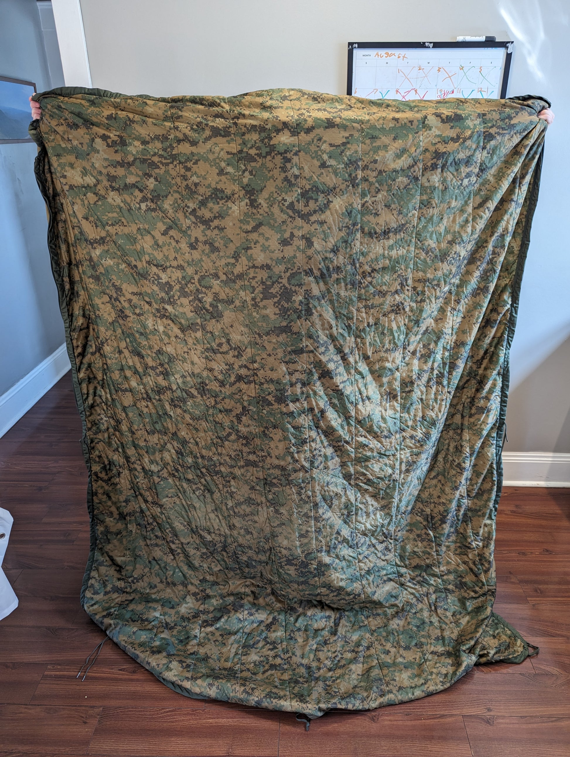 A full-size U.S. Military Surplus Poncho Liner (Woobie) in MARPAT digital camouflage, held up vertically by two people to demonstrate its full dimensions and quilted pattern.