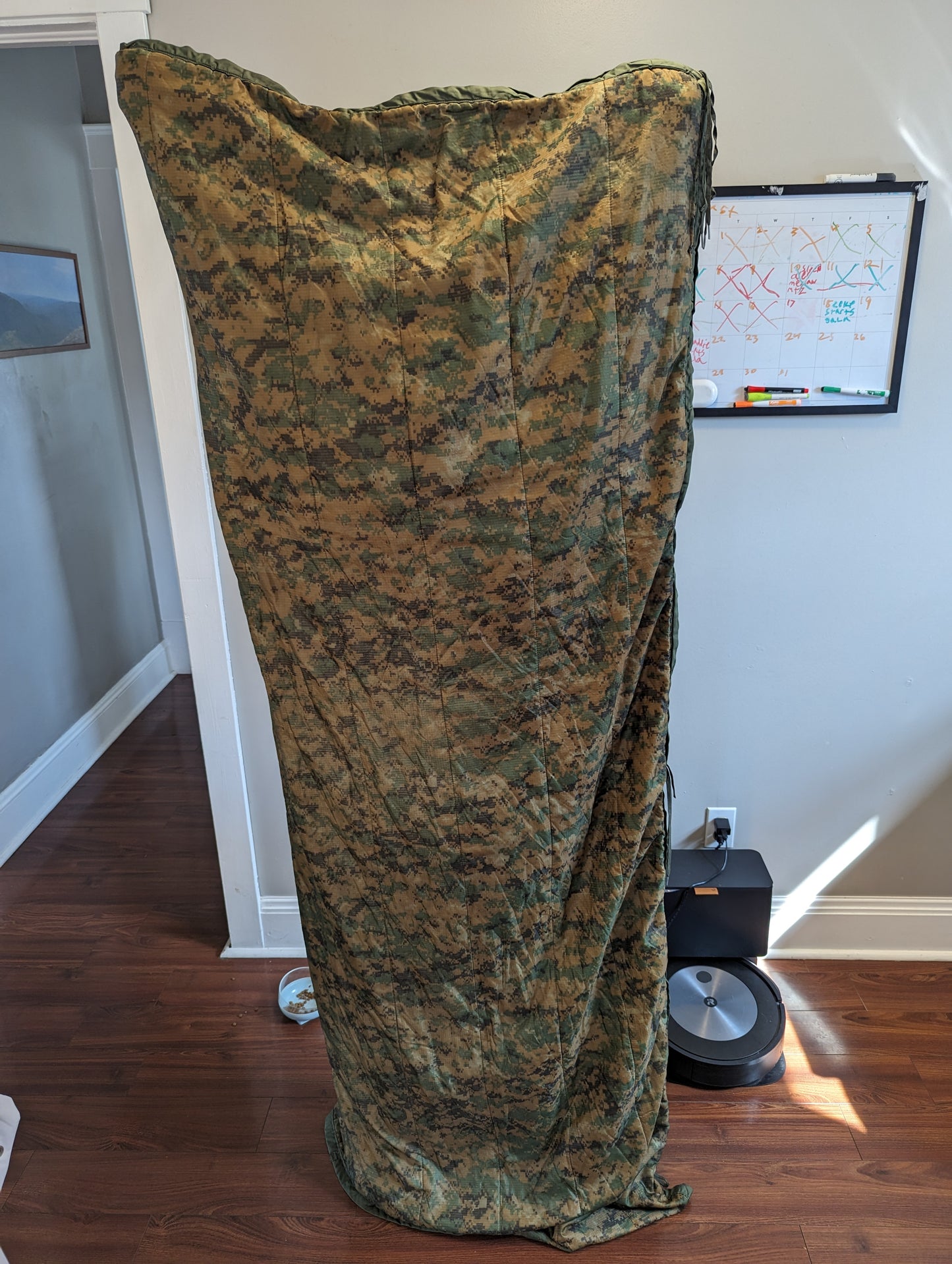 A U.S. Military Surplus Poncho Liner (Woobie) in MARPAT digital camouflage, rolled tightly into a compact cylinder and standing upright on a wooden floor, showcasing the quilted texture.