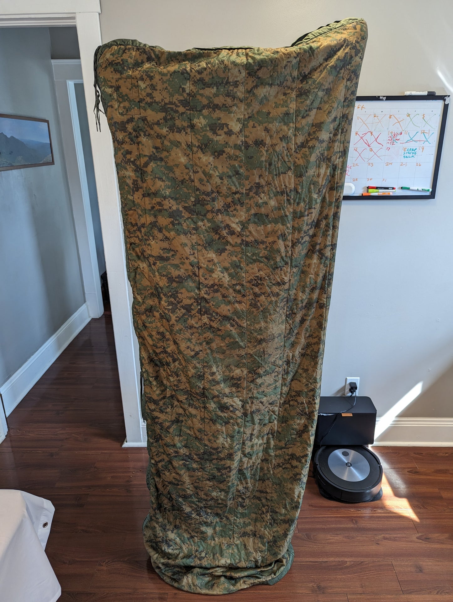 A U.S. Military Surplus Poncho Liner (Woobie) in MARPAT digital camouflage, rolled vertically and standing upright on a wooden floor, showcasing the heavy quilted pattern.