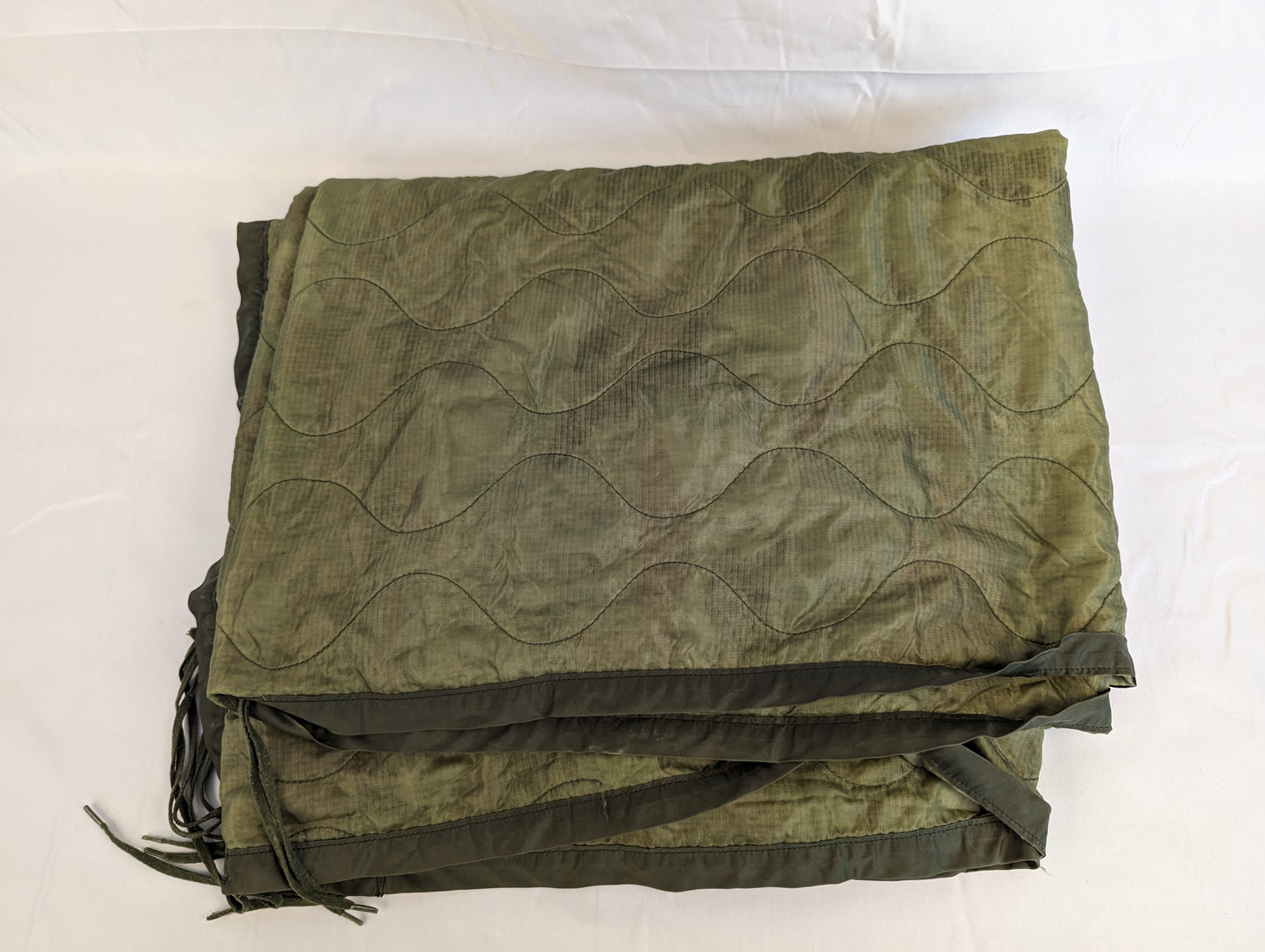 A U.S. Military Surplus Poncho Liner (Woobie) in solid olive green, folded flat, displaying the wavy quilted stitch pattern, dark binding, and attachment tie cords on a white background.