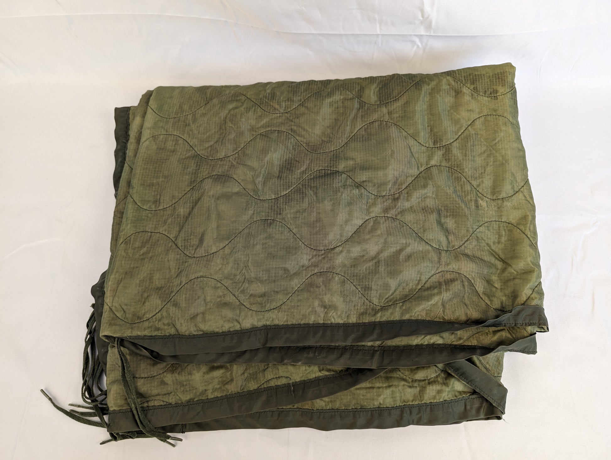 A U.S. Military Surplus Poncho Liner (Woobie) in solid olive green, folded flat, displaying the wavy quilted stitch pattern, dark binding, and attachment tie cords on a white background.