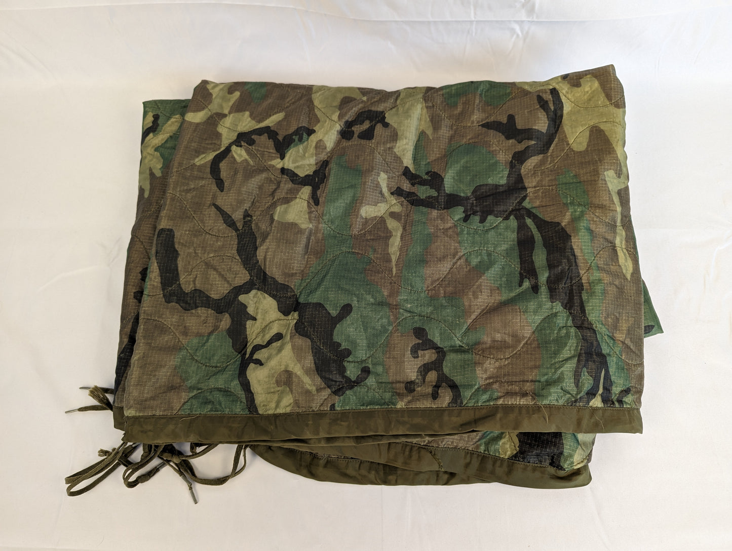 A U.S. Military Surplus Poncho Liner (Woobie) in Woodland camouflage, laid flat and folded to display the quilted diamond pattern, dark green edge binding, and attachment tie cords.