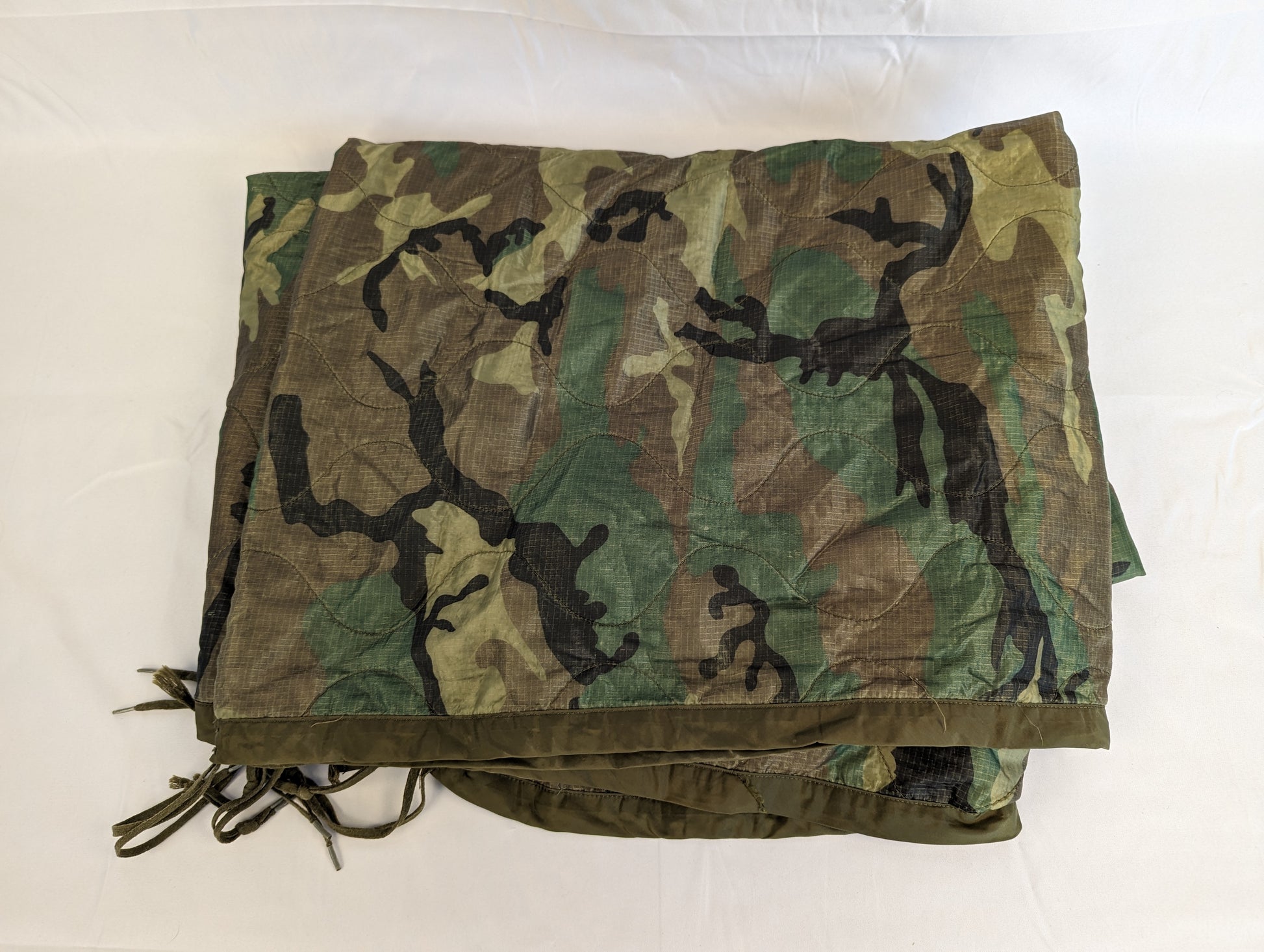 A U.S. Military Surplus Poncho Liner (Woobie) in Woodland camouflage, laid flat and folded to display the quilted diamond pattern, dark green edge binding, and attachment tie cords.