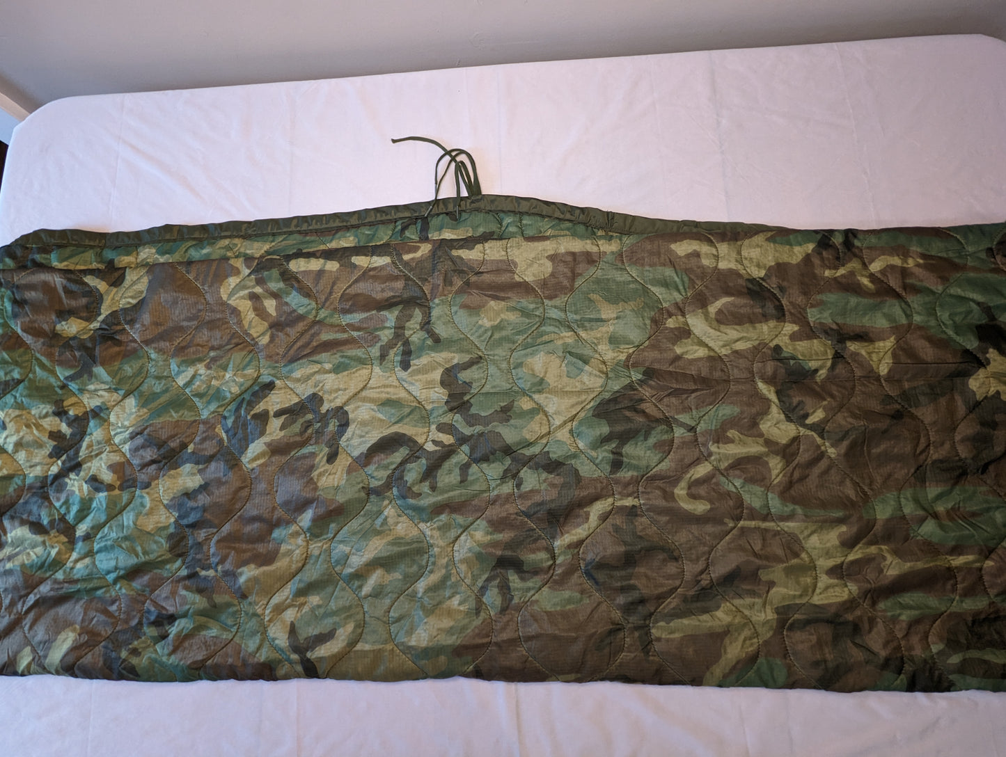 A U.S. Military Surplus Poncho Liner (Woobie) in Woodland camouflage, laid flat and folded horizontally, displaying the diamond quilted pattern, dark green edge binding, and attachment tie cords.