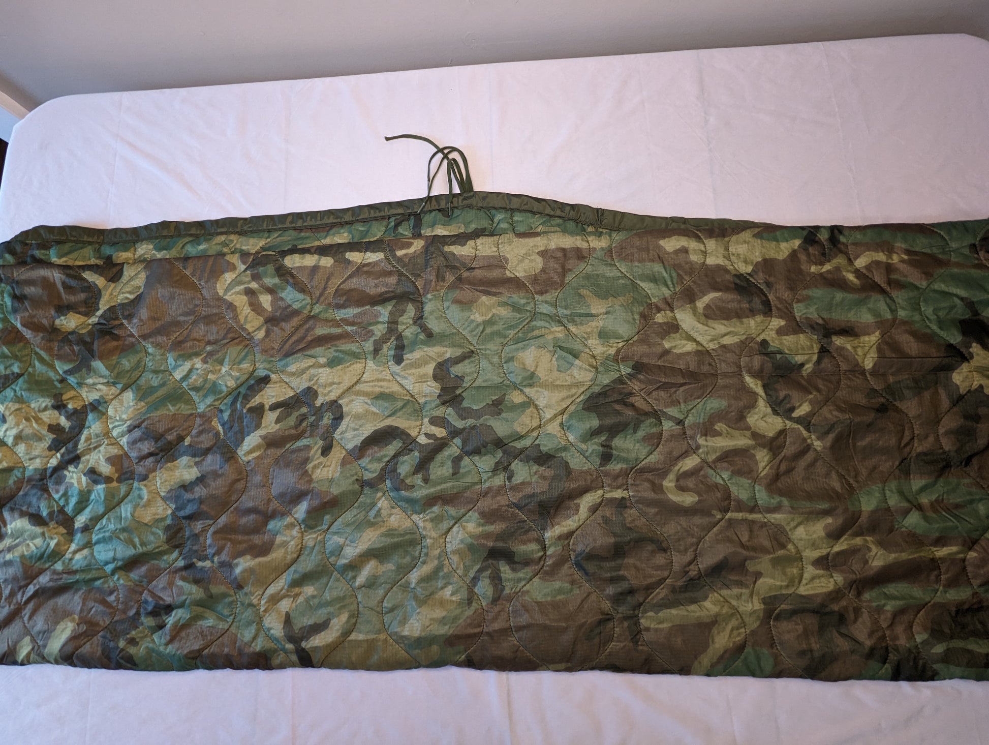 A U.S. Military Surplus Poncho Liner (Woobie) in Woodland camouflage, laid flat and folded horizontally, displaying the diamond quilted pattern, dark green edge binding, and attachment tie cords.