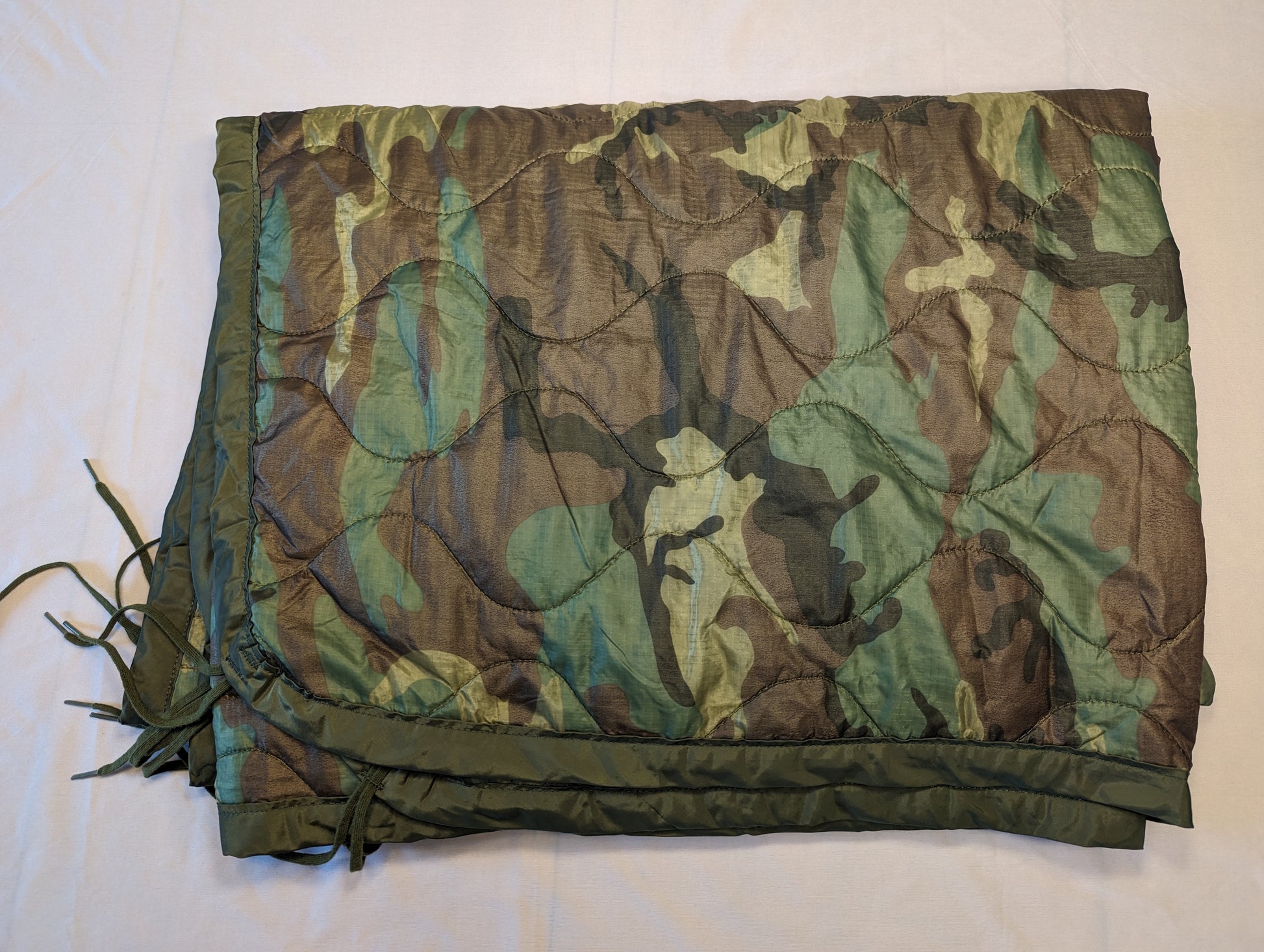 A full view of a U.S. Military Surplus Poncho Liner (Woobie) in Woodland camouflage, laid flat, displaying the diamond quilted pattern, dark green edge binding, and attachment tie cords.