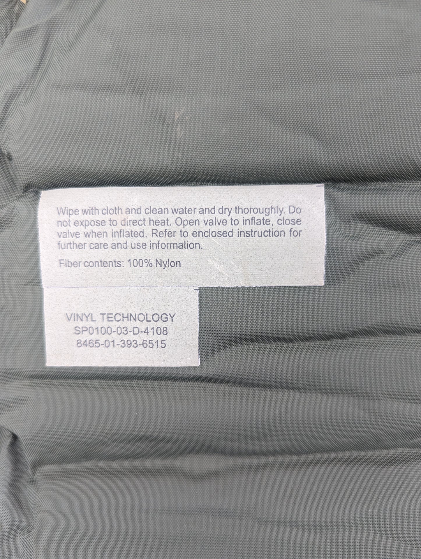 A close-up of the printed label on a U.S. Military self-inflating sleeping mat manufactured by Vinyl Technology, displaying usage instructions, contract number SP0100-03-D-4108, and NSN 8465-01-393-6515.