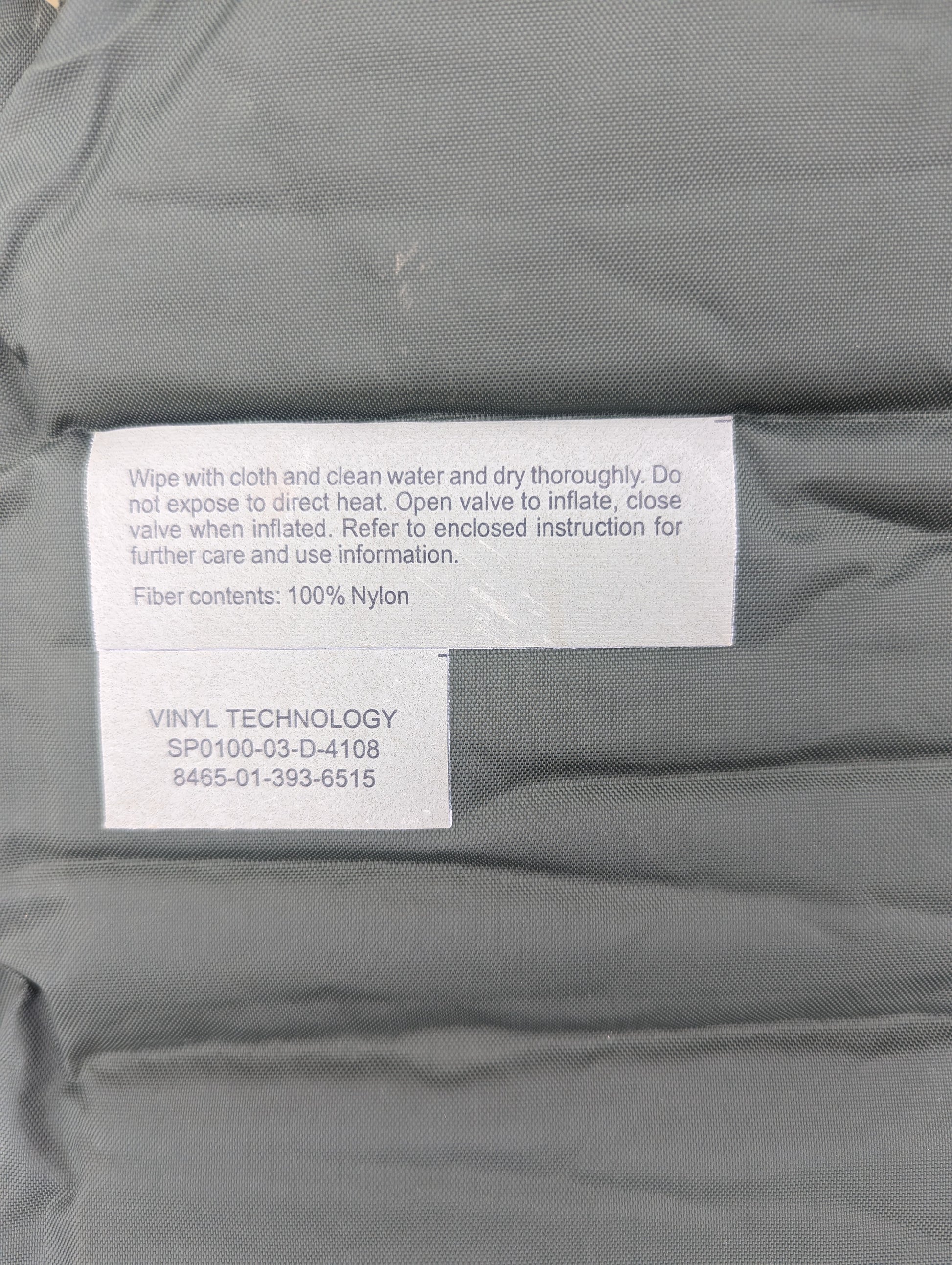 A close-up of the printed label on a U.S. Military self-inflating sleeping mat manufactured by Vinyl Technology, displaying usage instructions, contract number SP0100-03-D-4108, and NSN 8465-01-393-6515.