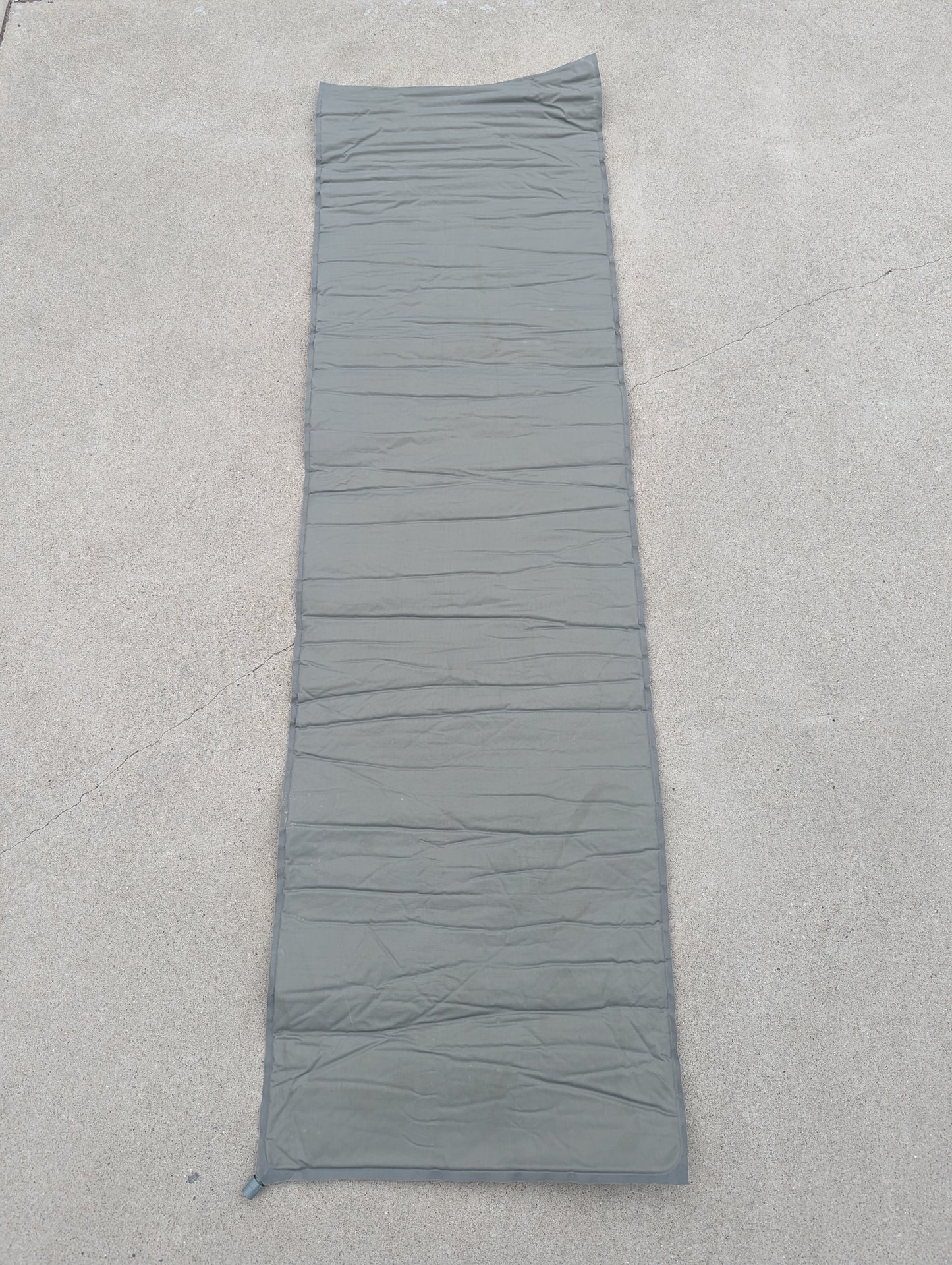 A full-length U.S. Military Surplus self-inflating sleeping mat in foliage green, laid flat on a concrete surface, highlighting the horizontal ribbed pattern and corner valve.