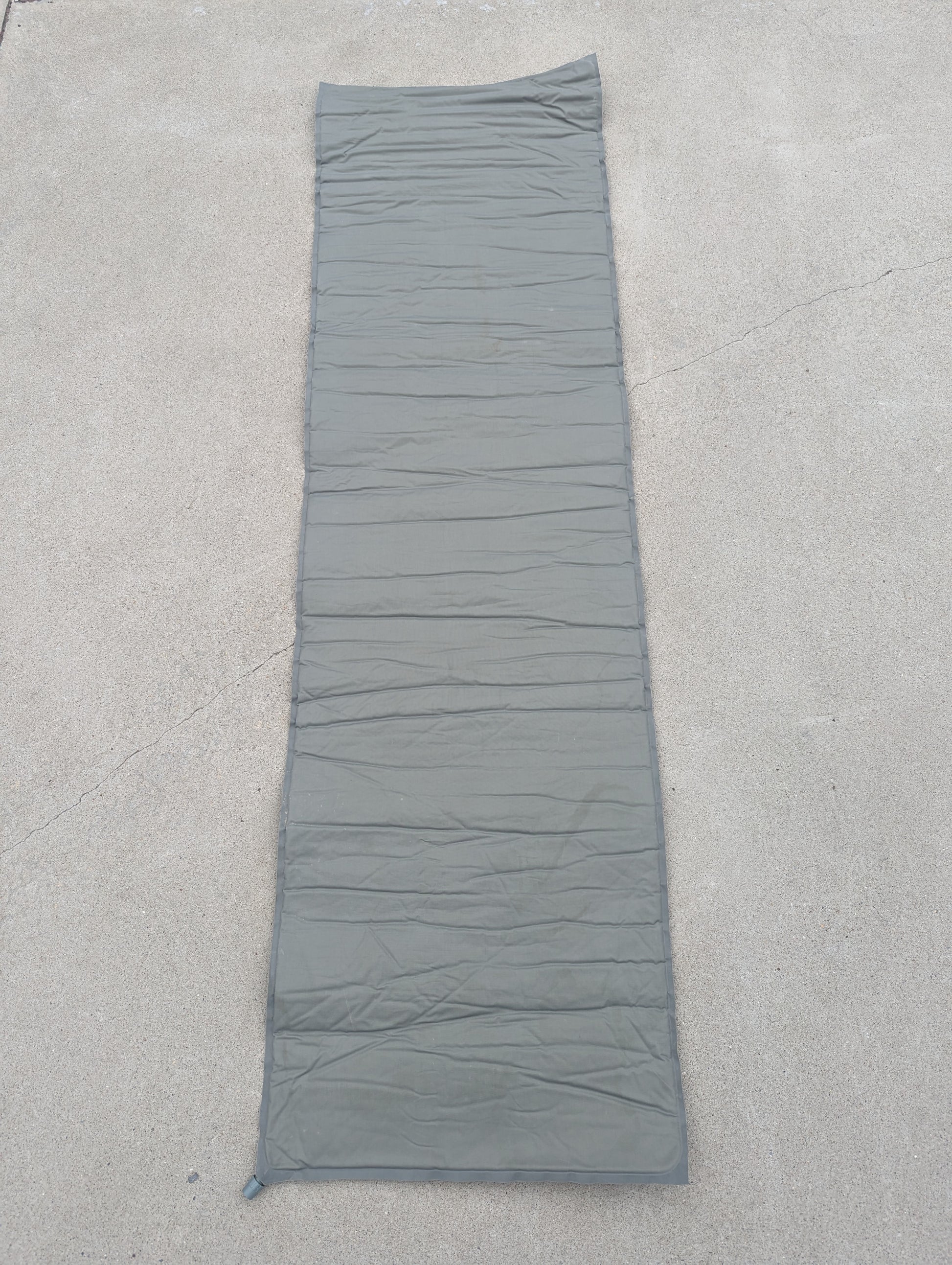 A full-length U.S. Military Surplus self-inflating sleeping mat in foliage green, laid flat on a concrete surface, highlighting the horizontal ribbed pattern and corner valve.