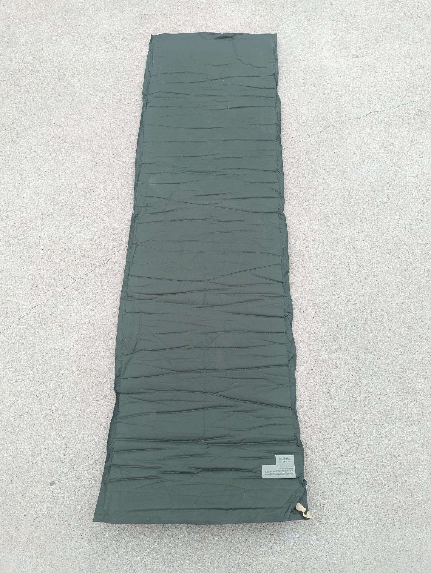 A full-length overhead view of a U.S. Military Surplus self-inflating sleeping mat in foliage green, laid flat on a concrete surface, highlighting the horizontal ribbed texture and corner valve.