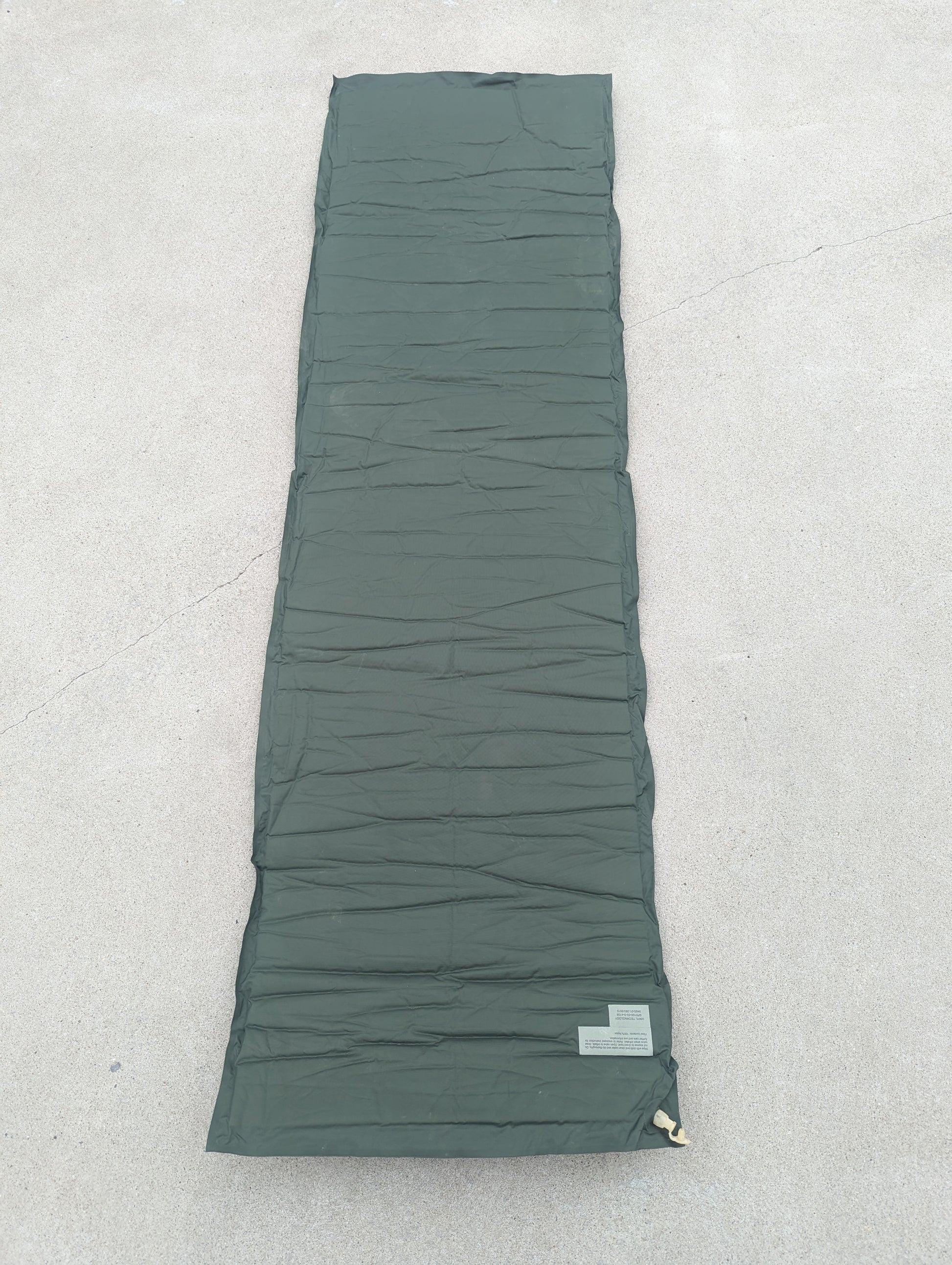 A full-length overhead view of a U.S. Military Surplus self-inflating sleeping mat in foliage green, laid flat on a concrete surface, highlighting the horizontal ribbed texture and corner valve.